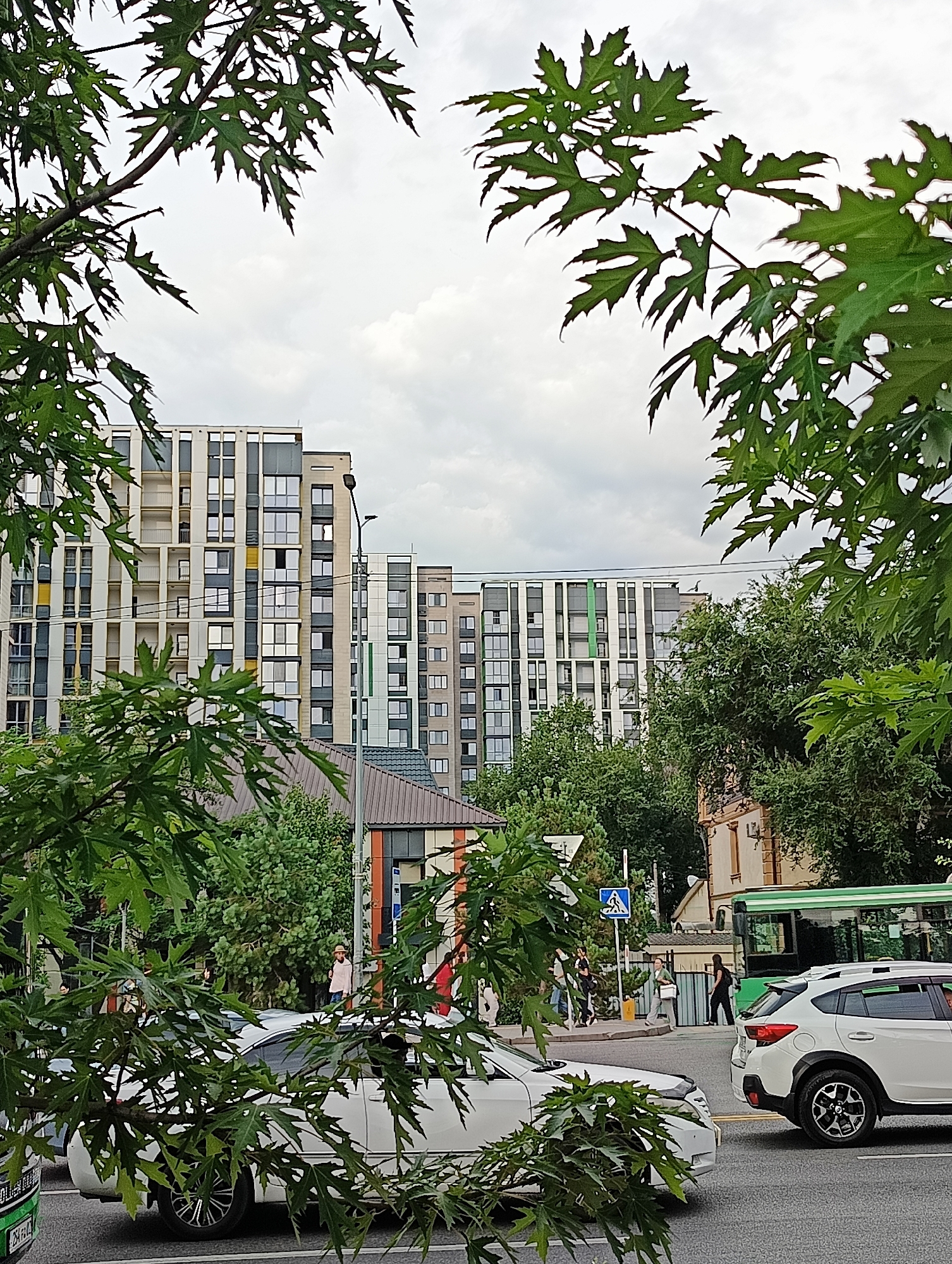 The image depicts a street scene in Almaty, Kazakhstan, framed by lush green foliage in the foreground. Modern, multi-story apartment buildings with a mix of white, grey, and accent colors dominate the background. The architecture suggests a contemporary urban environment.
In the middle ground, a green bus is visible, along with a white SUV and a white sedan on the road. Several people are walking on a sidewalk near a pedestrian crossing marked by a blue sign with a white pedestrian symbol. One person is wearing a straw hat and a light-colored top, while others are dressed casually in outdoor attire.
The sky is overcast, indicating a cloudy day. The presence of trees and the overall lushness of the foliage suggest it is likely daytime, possibly late spring or summer. The scene appears to be a typical urban street with residential buildings, traffic, and pedestrians. There is no discernible text that is legible enough to be transcribed. The mood of the image is neutral, depicting an ordinary day in the city.
