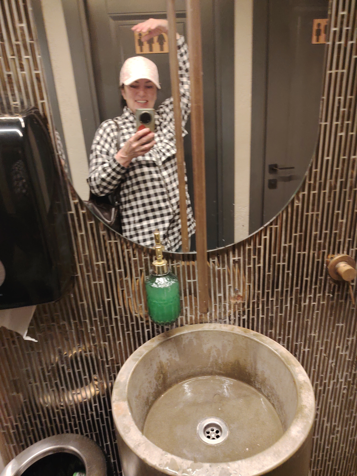 This image depicts a selfie taken in a public restroom mirror in Minsk, Belarus. A female subject, wearing a light pink baseball cap and a black-and-white checkered long-sleeved shirt, is smiling at her reflection while holding a smartphone with a gold-colored case to take the picture. Her right arm is raised to hold the phone, while her left arm is bent. The large, oval-shaped mirror reflects the subject and the surrounding bathroom elements. The wall surrounding the mirror and behind the sink features a distinctive, textured design composed of numerous narrow, vertically and horizontally arranged brown-toned tiles in a staggered pattern. In the lower foreground, a round, light beige, concrete-like pedestal sink is visible, containing a small amount of residual water in its basin and featuring a metallic drain. A green, textured glass soap dispenser with a brass pump top is reflected to the left of the sink. On the far left, a black wall-mounted paper towel dispenser with a visible white paper towel is also reflected. In the background reflection, a dark grey door with a black handle and a light-colored vertical trim is seen, along with a small, rectangular wooden sign depicting universal male and female restroom symbols.