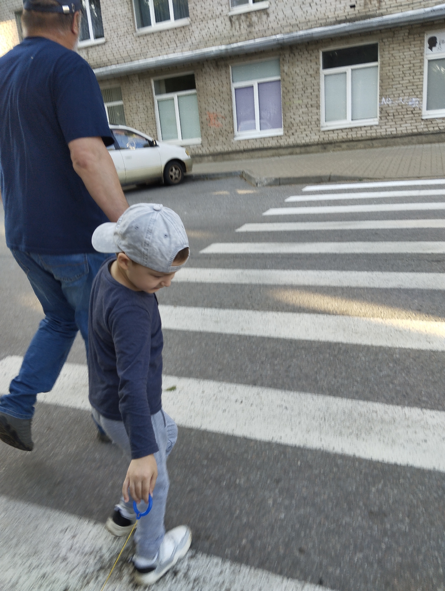 In Kingisepp, Russia, on a street with a white-lined pedestrian crossing, a child wearing a grey baseball cap and a dark blue long-sleeved shirt walks across the street. The child is holding a blue ring with a yellow string attached, possibly part of a toy. Beside the child, an adult wearing a dark blue t-shirt and blue jeans is also walking across the pedestrian crossing. The adult is partially obscured by the child. In the background, a light-colored car is parked on the side of the street, and a brick building with several windows is visible. The scene appears to be captured during daylight hours.