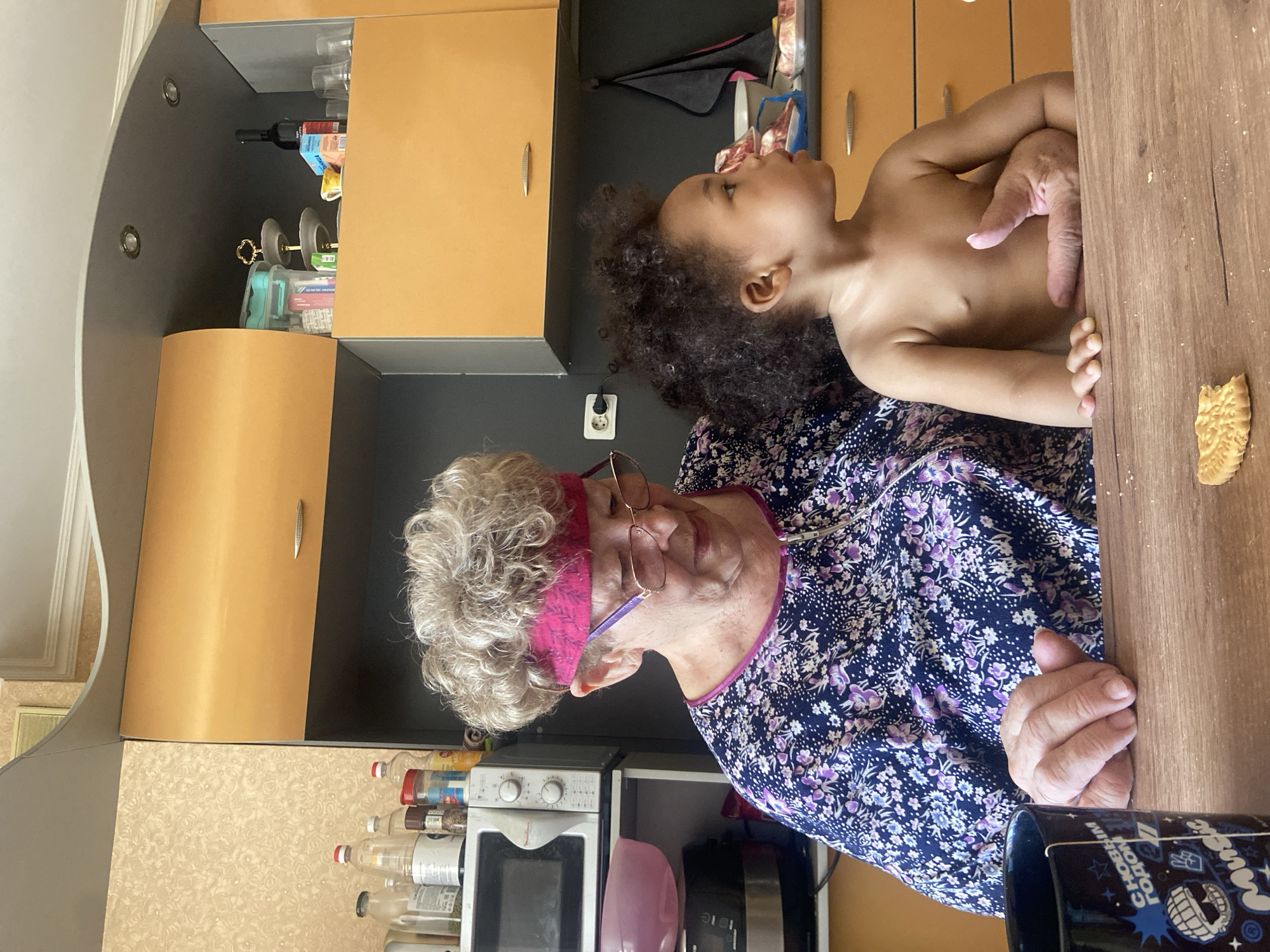 An elderly woman with curly grey hair and a pink headband sits at a wooden table in a kitchen, holding a shirtless toddler with dark curly hair. The woman is wearing a floral patterned top and glasses. The toddler is looking upwards. A cookie is on the table. Behind them are kitchen cabinets in a combination of light wood and dark grey. A microwave and various containers are visible on the countertop. A black mug with cartoonish white designs is partially visible at the lower right corner. The location is Kazan, Russia.