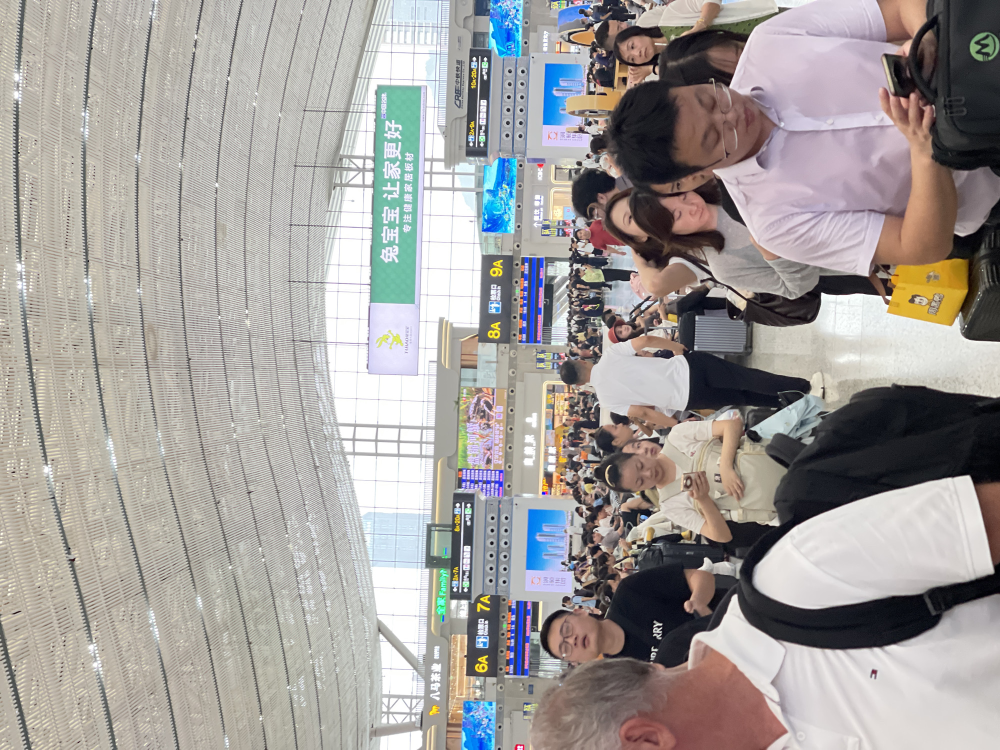 This media file captures a busy scene inside a modern transportation hub, likely a train station or airport in Longhua District, China, judging by the visible text. The image is oriented vertically, with the ceiling structure dominating the upper portion and a crowd of people filling the lower half.
The ceiling is a prominent architectural feature, made of a white, latticed material that curves upwards, suggesting a large, open space. Large digital display boards are visible throughout the scene, showing train or flight information, advertisements, and time schedules, indicated by text like "8A", "9A", "6A", and "2A-7A". One prominent green banner displays Chinese characters, likely an advertisement for "兔宝宝" (Rabbit Baby), promoting healthy home building materials. Other visible text includes "ICBC" (Industrial and Commercial Bank of China) and "CRF中铁快运" (China Railway Express).
A large crowd of people are seen waiting, many seated on chairs or on the floor, suggesting a period of delay or high passenger volume. They appear to be a mix of ages, with individuals looking at their phones, talking, or resting. The atmosphere appears to be one of typical travel congestion, with a sense of hurriedness mixed with patience. The lighting suggests it's daytime, with natural light filtering in from above. Some individuals are carrying luggage, further reinforcing the travel context. The overall impression is a snapshot of daily life and transit in a bustling urban environment.