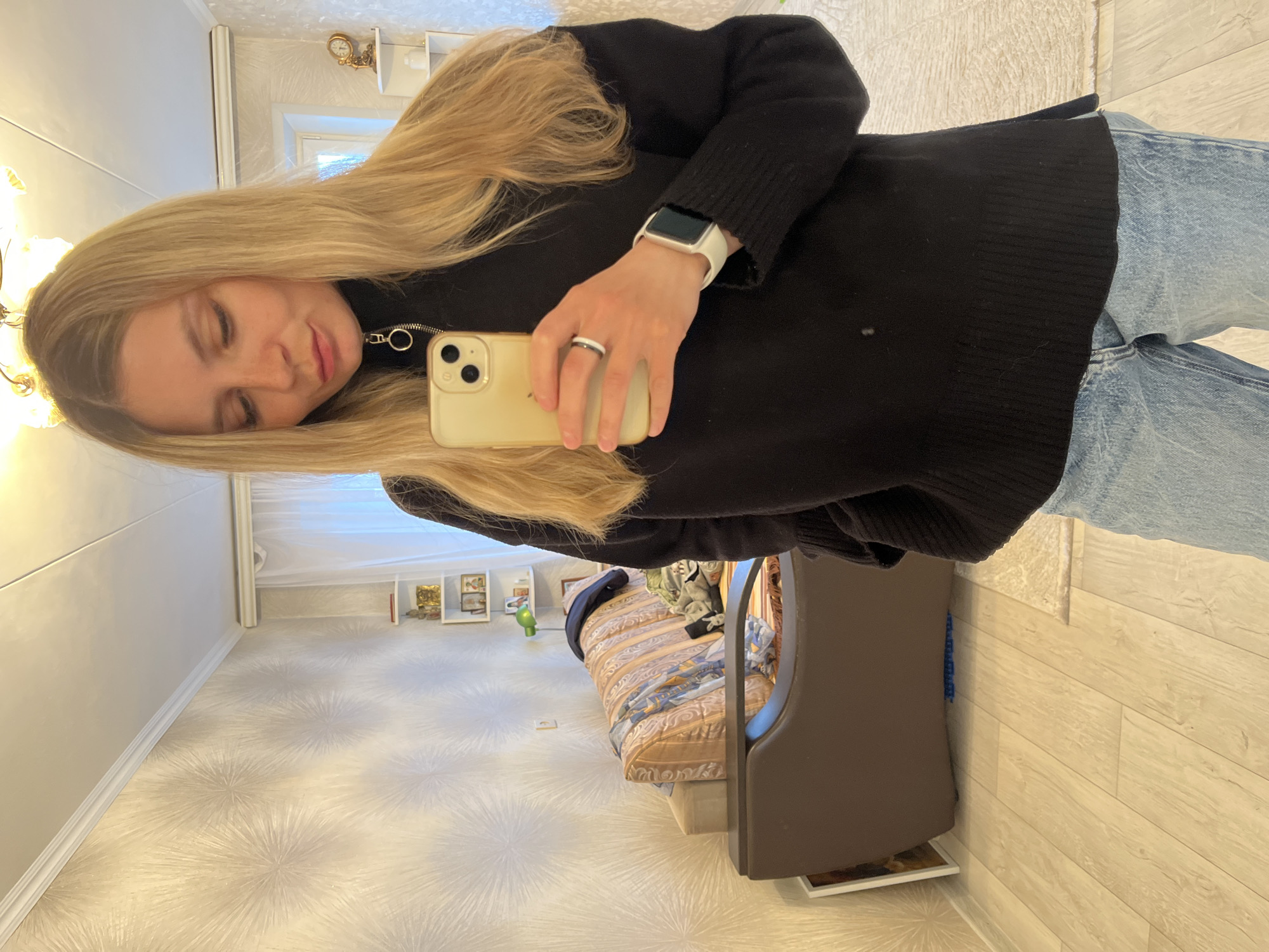 A fair-skinned female with long blonde hair is taking a selfie indoors. She is wearing a black sweater with a zipper detail, blue jeans, a white smartwatch, and a ring. She is holding a gold-colored iPhone. The background reveals a room with floral wallpaper, a bed with patterned bedding, a brown sofa, and a cabinet with decorative items. The location is Chaikovsky, Russia.