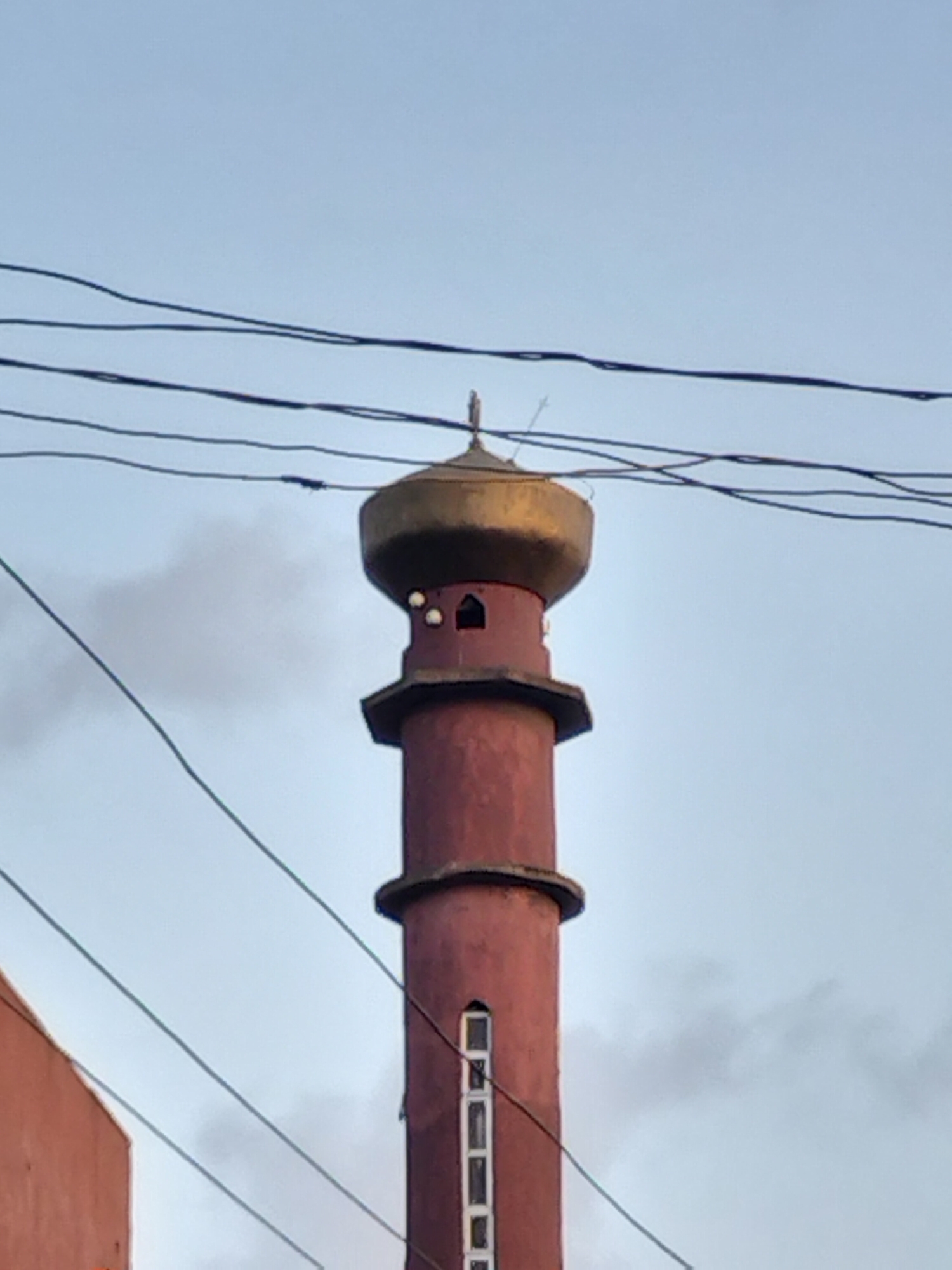 A minaret, possibly part of a mosque, is the central subject of the image. The minaret is a multi-tiered structure, primarily red-brown in color. It features a bulbous, golden dome at its apex, topped with a small spire. The tower has at least two ringed sections and a series of small, vertically stacked windows. Power lines cut across the frame in the foreground, obscuring some details of the minaret. The sky behind is a muted blue-grey. The lower left corner of the image shows a small portion of a building, similarly colored to the minaret. The location is suggested to be in Lagos, Nigeria.