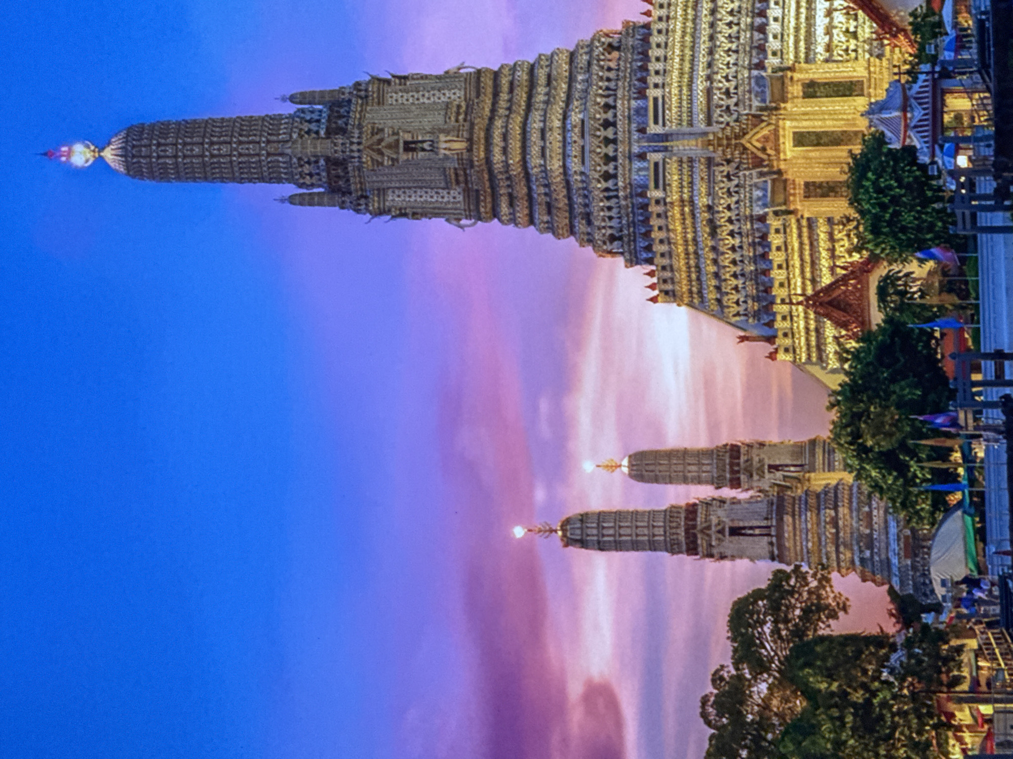 The image captures a twilight view of Wat Arun, also known as the Temple of Dawn, in Bangkok, Thailand. The central focus is the temple's prang, a towering spire adorned with intricate porcelain mosaics and topped with a finial that glows brightly. To the left of the main prang, two smaller spires stand, also illuminated. The sky transitions from a deep blue at the top to hues of purple and pink, with soft, wispy clouds. Lush green trees frame the bottom left and right of the scene, with some foliage obscuring parts of the temple's base. In the foreground, a waterfront area is visible with structures that appear to be buildings, piers, and boats, suggesting it is situated along a river. Small flags are present on the waterfront. The lighting indicates it is either sunrise or sunset, with the temple and its spires illuminated. The city of Bellheim, Germany is not depicted in this image.