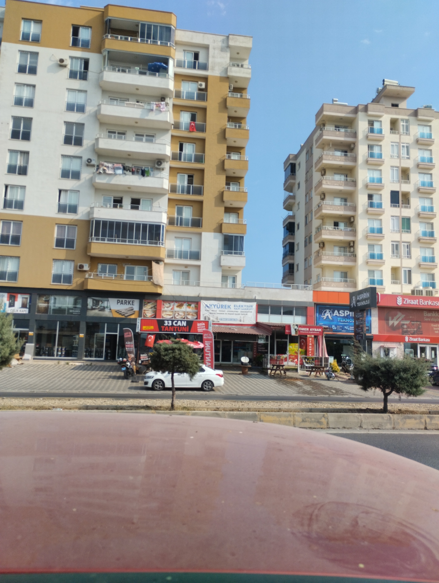 In the city of Erdemli, Turkey, two tall, modern apartment buildings stand under a clear blue sky. The building on the left is primarily white with yellow accents, featuring multiple balconies with glass railings and some with enclosed glass rooms. On the right, another apartment building is predominantly cream-colored with light blue glass balconies. Both buildings have visible air conditioning units and windows.
At the base of the buildings, a commercial area is visible. Storefronts display various signs, including "PARKE," "33 CAN TANTUNI," "ASPIRIN TEKNOLOJI," and "Ziraat Bankasi." Several shops appear to be open, with awnings and displays.
In front of the shops, a paved walkway is lined with trees and street furniture, including picnic tables and benches. A white sedan is parked on the street, and a motorcycle is parked on the walkway. The street is paved and appears to be a busy thoroughfare. The foreground of the image shows the hood of a red car, obscuring the lower part of the scene. The overall impression is a contemporary urban environment in Turkey.