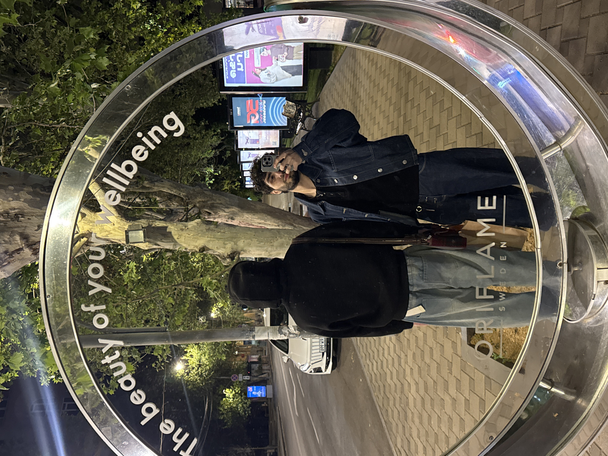 A man with dark hair and a beard is taking a selfie with his phone in a reflective, circular public art installation, located in Yerevan, Armenia. The installation reads "The beauty of your wellbeing" around the perimeter. The reflection also shows a person wearing a dark hoodie and light-colored pants, standing behind the man taking the photo. The reflection shows the street scene with buildings, trees, street lights, and billboards in the background. The word "ORIFLAME SWEDEN" is visible in the reflection on the lower right side of the circle. The street contains a car with the license plate "8९९ ня".