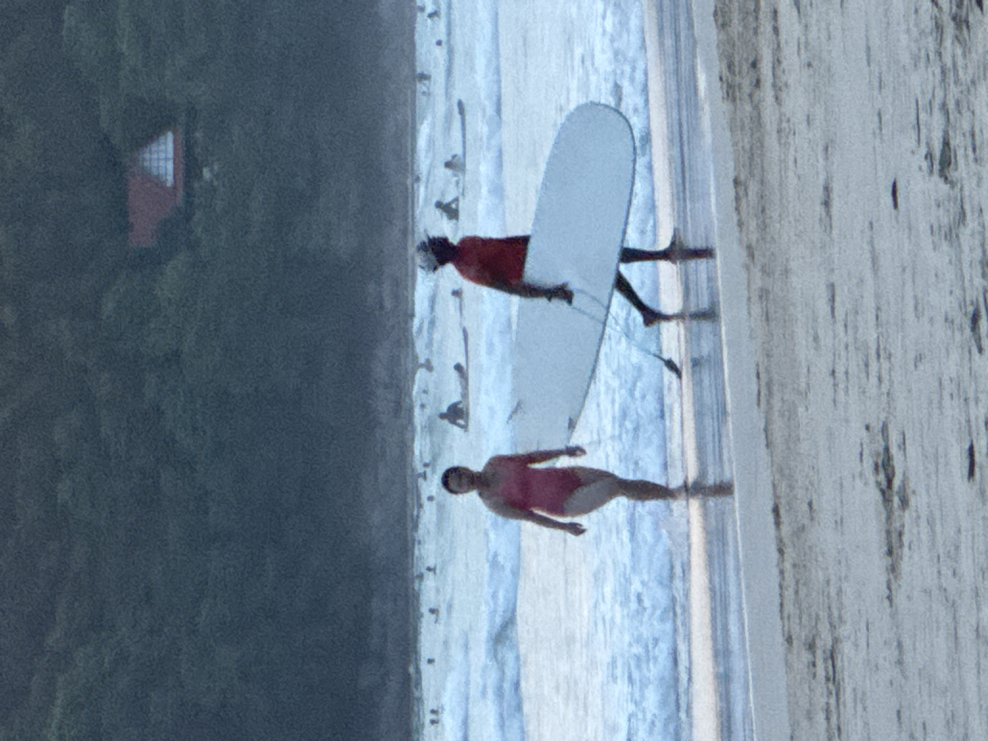 In Karon, Thailand, a beach scene unfolds with a woman in a pink swimsuit walking on wet sand near the water's edge. To her right, a man in a red shirt and cap carries a white surfboard. The ocean is moderately wavy with several other surfers visible in the distance. Behind the beach, a dense, dark green forested hillside rises, with a red-roofed building with a triangular window peeking through the foliage. The sky is overcast and dark.