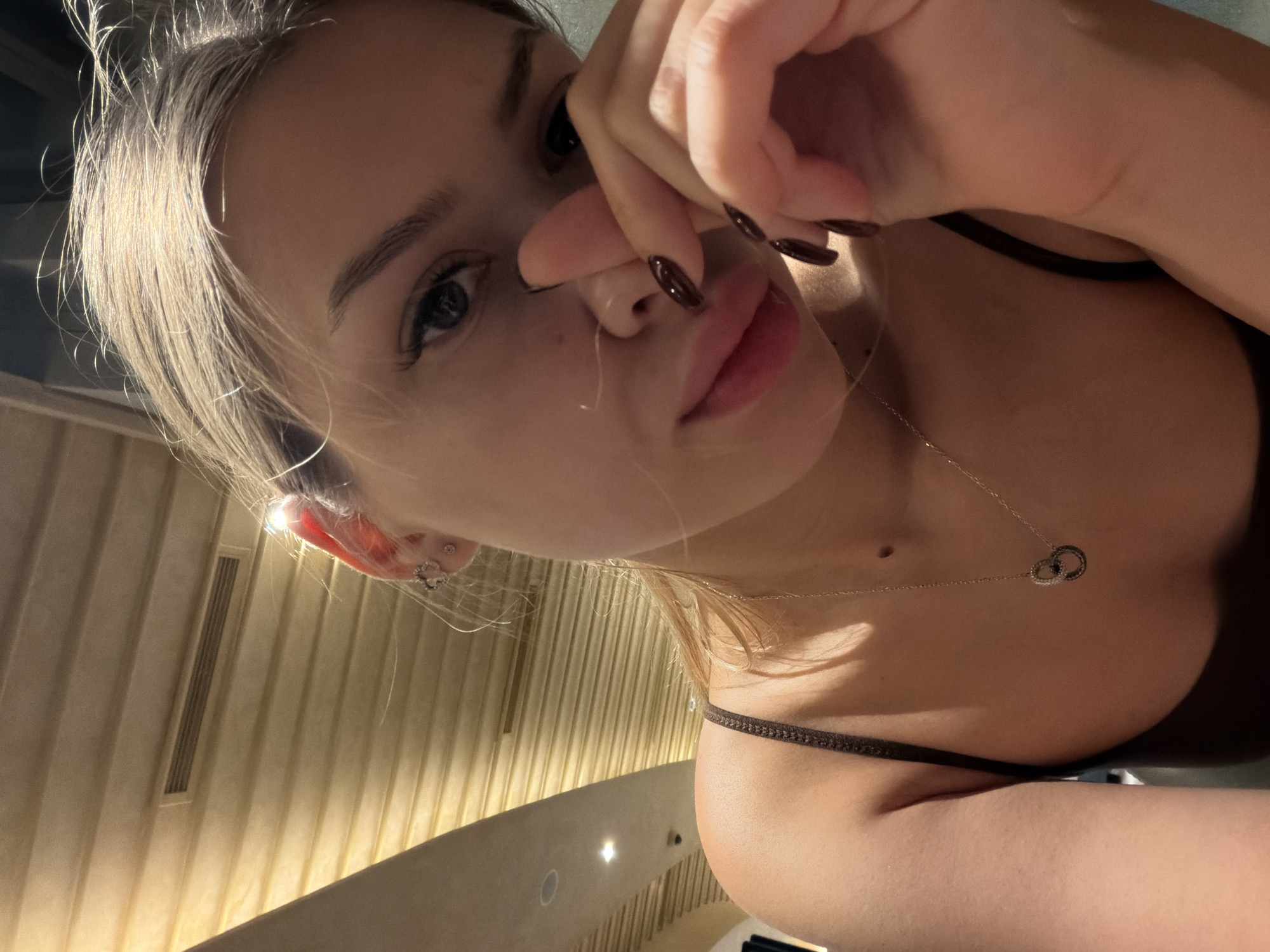 The image shows a close-up selfie of a young Caucasian woman. She has blonde hair and appears to be indoors, possibly in a restaurant or a similar establishment, given the wood-paneled ceiling in the background. She is wearing a dark-colored top with a thin strap and a delicate silver necklace with a pendant. Her hand is raised, with her fingers touching her face near her nose. She has dark nail polish on her fingernails. The lighting suggests it might be evening or an interior setting with artificial light. There is no indication of the location being in the city of Đà Nẵng, Vietnam.