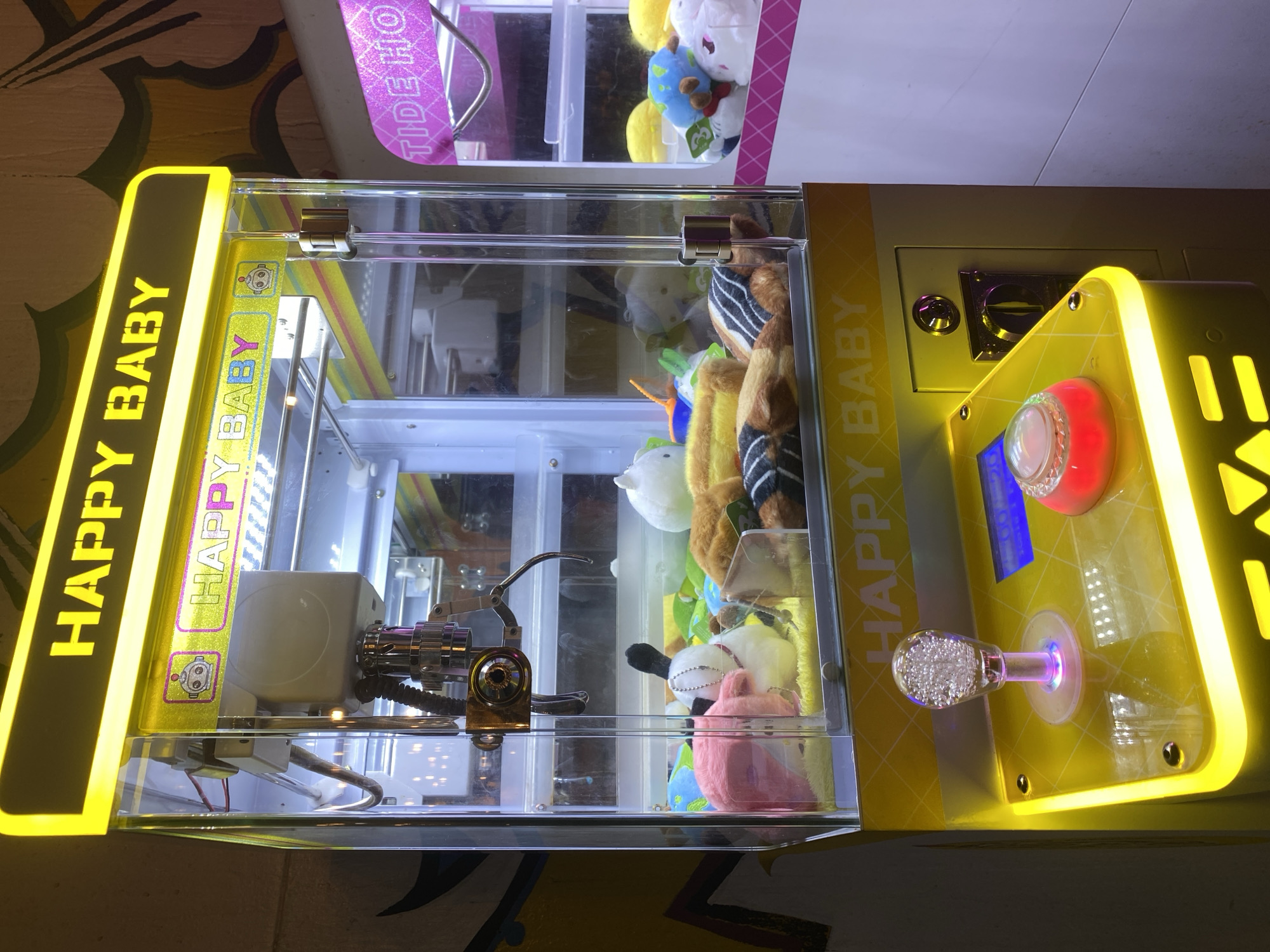 A close-up shot of a "Happy Baby" claw machine, filled with various plush toys, inside an entertainment establishment in Davao City, Philippines. The machine is brightly lit with yellow neon lights, and the control panel features a joystick with a clear, bubble-filled knob, a large red button, and a small blue screen. The claw mechanism is visible inside the glass enclosure, with several stuffed animals, including a pink one, a white one, and a striped one. Another claw machine labeled “Tide Ho" is partially visible above. The machine appears to be located indoors, with patterned wallpaper visible in the background.