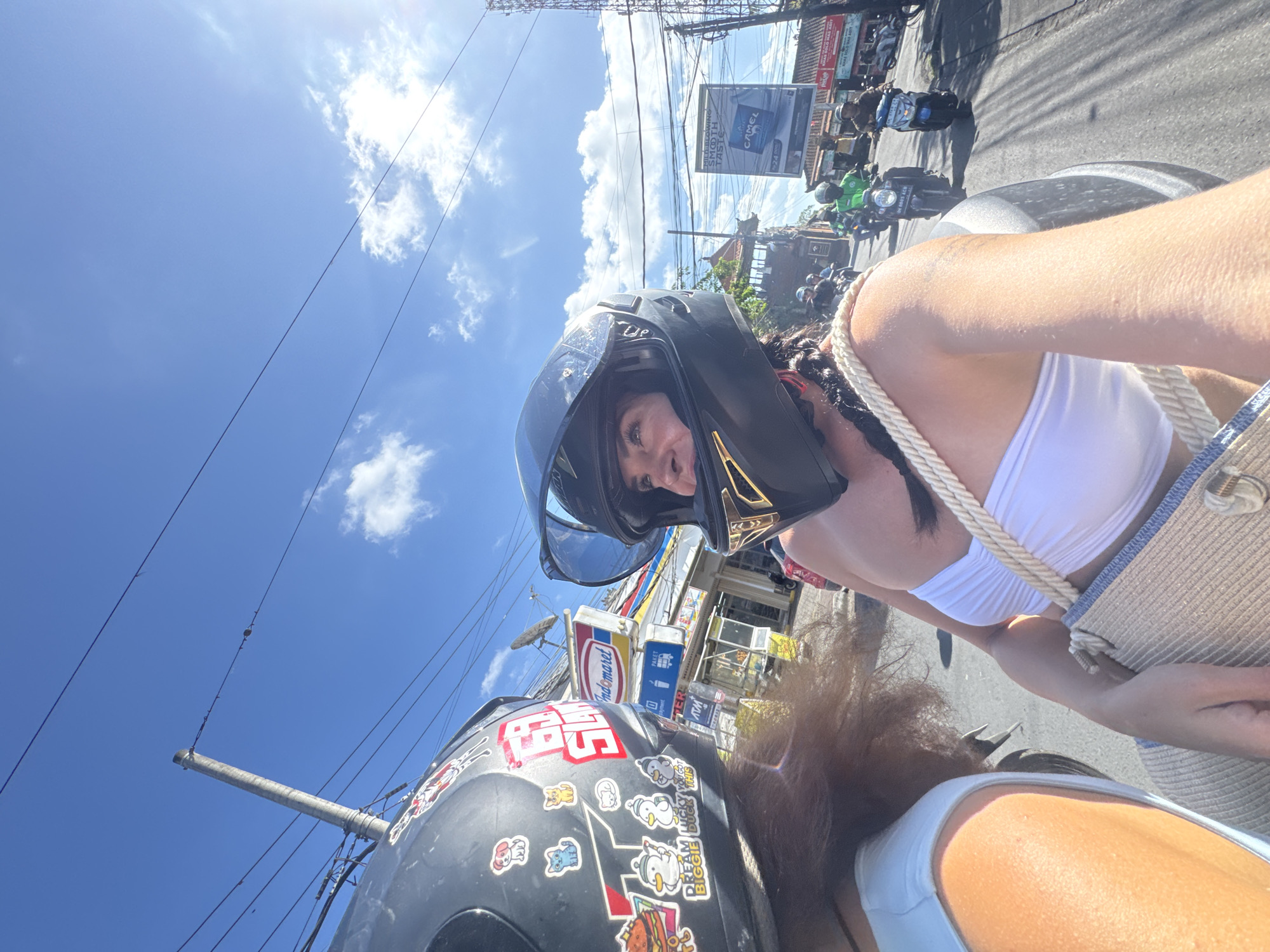 The image shows two people on a motorcycle in what appears to be a busy street in North Kuta, Indonesia. The subjects are a woman in the front wearing a black helmet with gold accents and a white bikini top, and another person in the back, whose face is not visible, also wearing a white top. The woman in front is looking at the camera with a smile. The scene is outdoors on a sunny day with a bright blue sky and some white clouds. Overhead wires and poles are visible, suggesting an urban or semi-urban environment.
Various objects are present in the scene. The motorcycle has a black helmet with stickers on it. In the background, there are shops and storefronts, including one with signs that read "Indomaret" and "ATM." Other motorcycles and people are visible on the street, indicating traffic and activity. A billboard with an advertisement for "Camel" cigarettes is also visible. The overall impression is one of a vibrant and bustling street in a tropical location.
The time of day appears to be daytime, given the bright sunlight and clear sky. The weather seems to be warm and sunny. The presence of shops, motorcycles, and overhead wires points to a developed area. The woman's smile and the overall scene suggest a casual and perhaps adventurous outing.