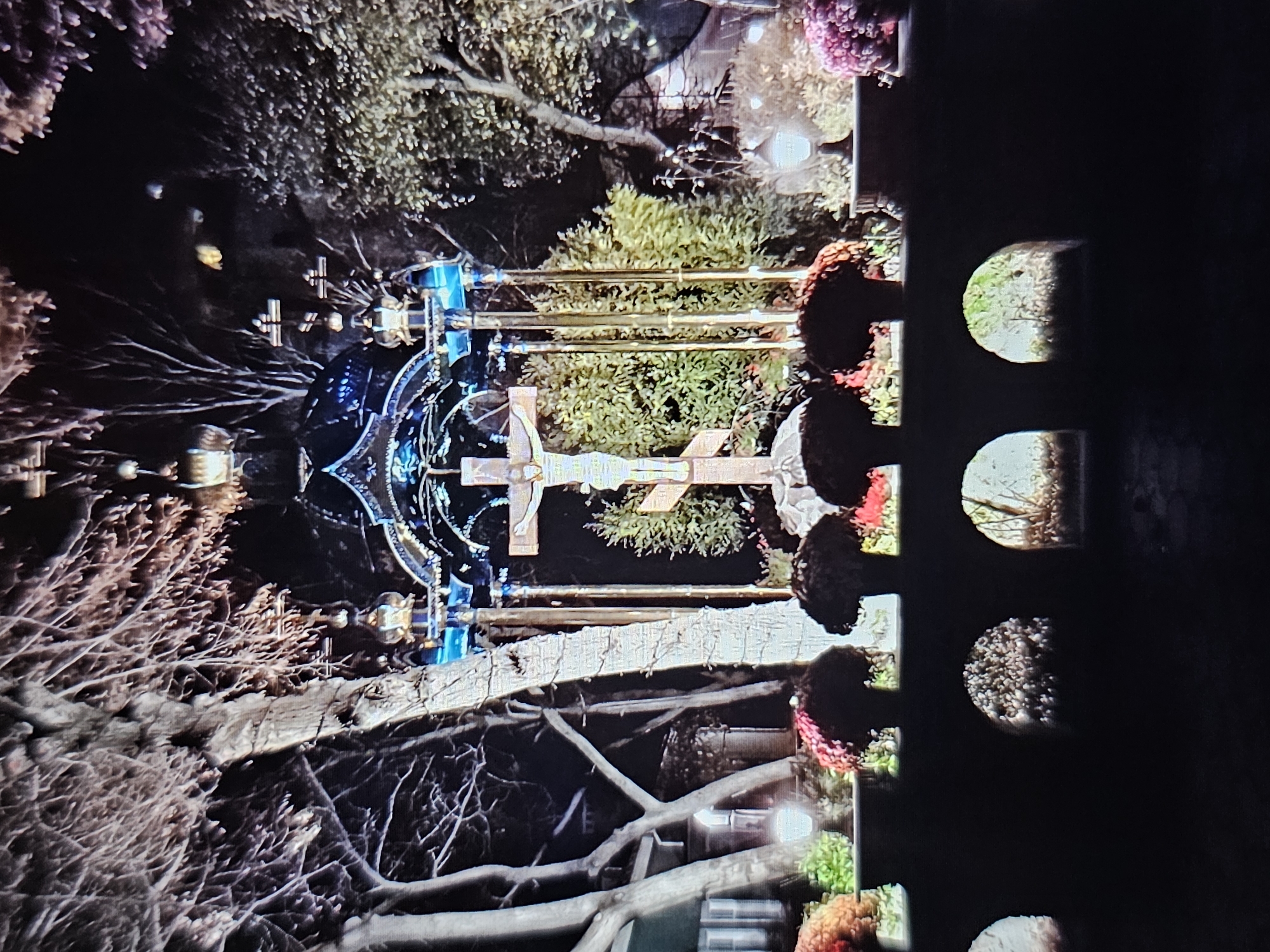 The image shows an outdoor nighttime scene at a cemetery in Amarillo, Texas. In the center, a large, ornate, multi-tiered structure with blue lights and gold accents stands over a wooden crucifix with the figure of Jesus. The crucifix is surrounded by lush green foliage. In front of the crucifix, there is a low wall with decorative arched openings below. On top of the wall, there are several arrangements of flowers in dark pots, including some that are red and deep pink. Two lampposts with glowing lights are visible on either side of the flower arrangements. The background features bare trees with intricate branches and some houses in the distance. The overall impression is one of solemnity and artistic illumination.