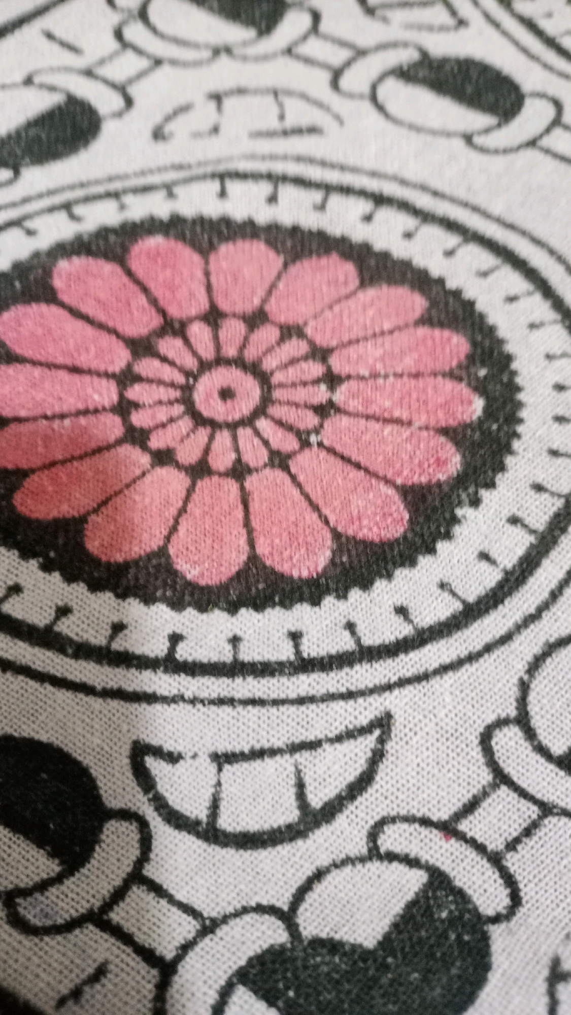 The image displays a close-up view of a fabric with a repeating pattern. The pattern features a central pink flower with black outlines and accents, surrounded by concentric circles with decorative elements. In the lower portion of the image, parts of black and white cartoon-like figures, possibly Mickey Mouse heads, are visible. The fabric appears to be white with black and pink print. The image does not provide any information about its location in the city of Deoghar, India, or any details about people, actions, or interactions.