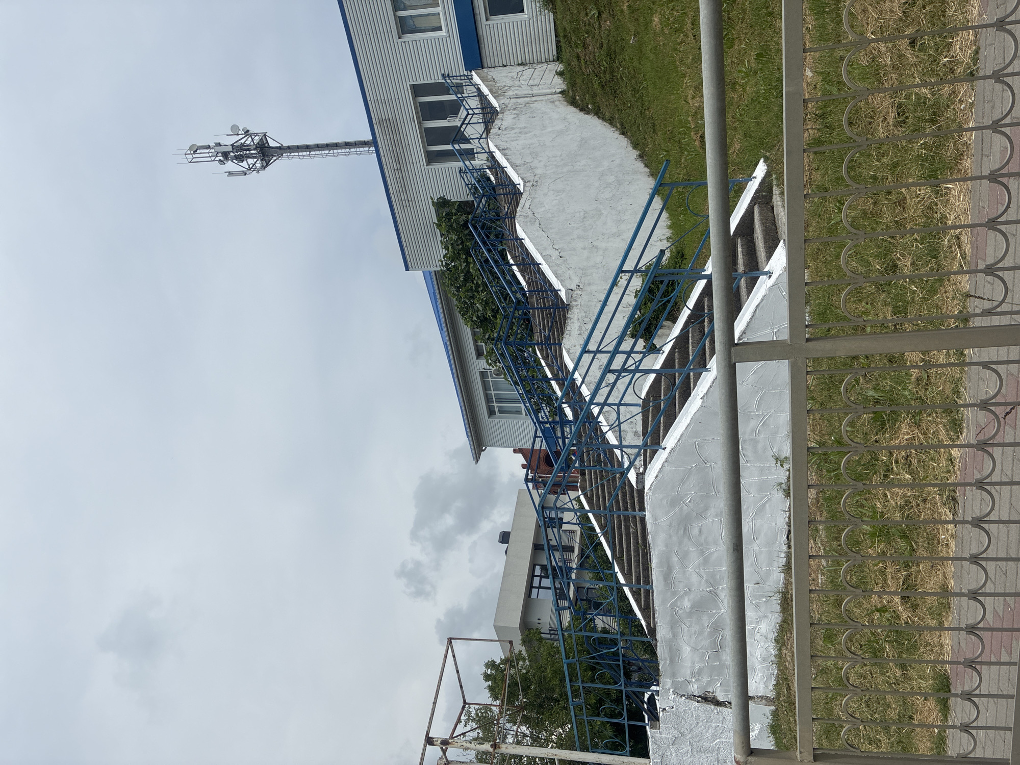 The image shows an outdoor scene with a long, white concrete staircase leading up a grassy hill. The staircase has blue railings and is bordered by green grass. At the top of the stairs, a modern, light-colored building with a blue roof is visible. A tall telecommunications tower with multiple antennas stands against a cloudy sky, positioned behind and to the right of the building. In the foreground, a grey metal fence with decorative arches runs horizontally across the bottom of the image, separating it from a paved area. The environment suggests a public space, possibly near a residential or commercial area in Arkhipo-Osipovka, Russia. There are no people or animals visible in the frame.