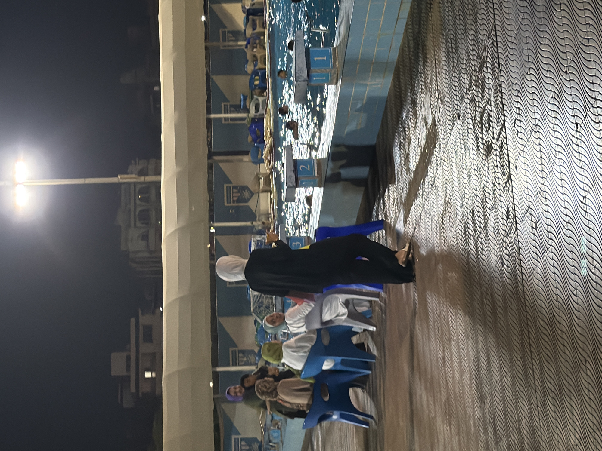 The image depicts an outdoor swimming pool area at night in the city of 6th of October, Egypt. In the foreground, the patterned tiled ground reflects light. To the left, a group of people are seated on blue and white chairs. Most of them appear to be women wearing headscarves, with one woman standing in a black abaya and headscarf holding a cup. In the background, the swimming pool is visible with several people in the water. There are diving blocks with numbers "1" and "2" visible on them. The area is illuminated by overhead lights. Buildings and a light pole are visible in the distance against the dark night sky.