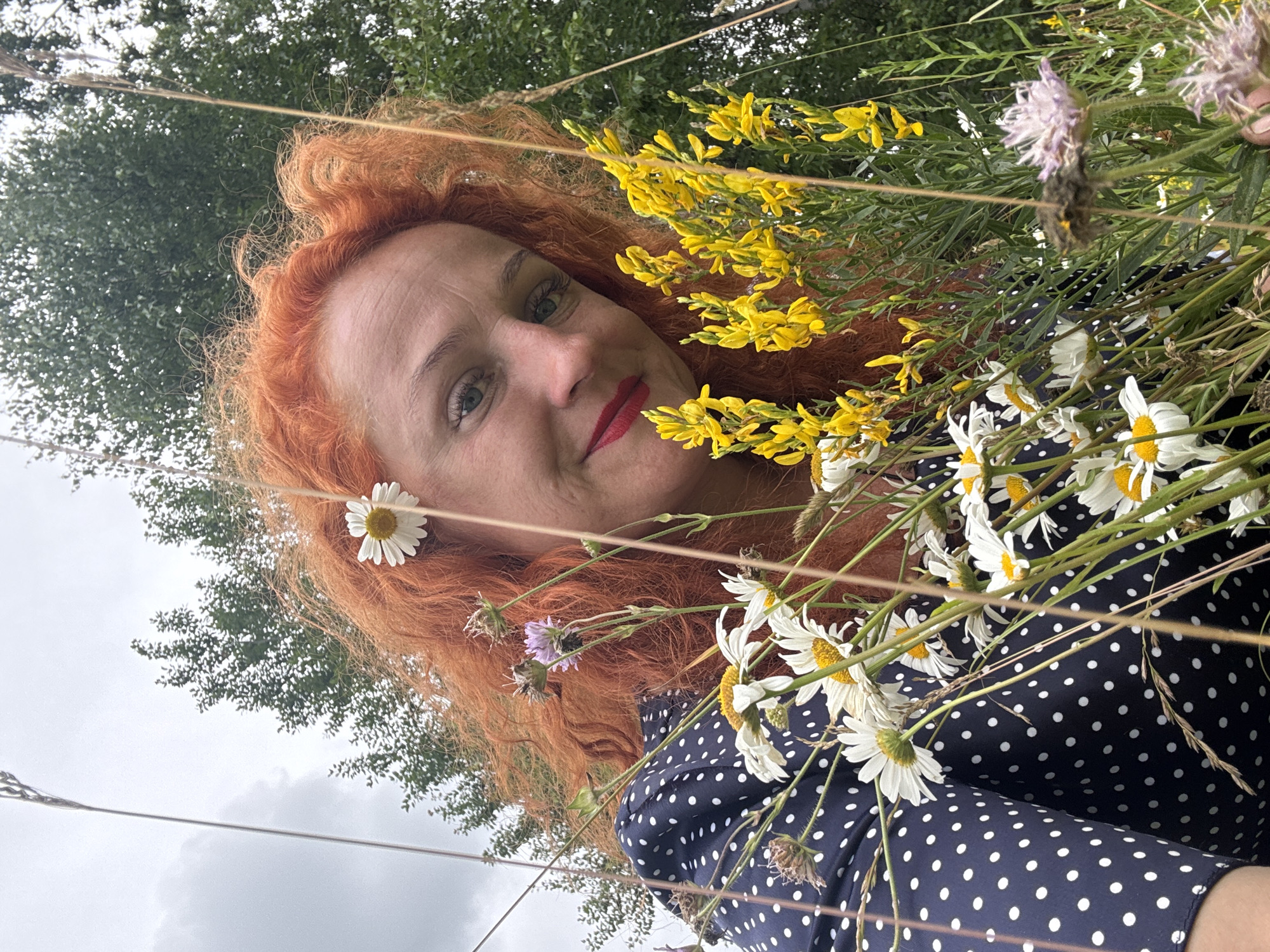 The image shows a woman with red, curly hair, her head tilted towards the camera, smiling and holding a bouquet of wild flowers. She is wearing a dark blue dress with white polka dots. She has red lipstick on. The bouquet consists of daisies, yellow broom flowers, and some other smaller purple and pink flowers. She is outdoors, with a background of green trees and a cloudy sky. The photographer is presumably close to the woman, as the perspective is from a low angle, looking up at her. There are no visible buildings or landmarks that would indicate a specific location within Privolzhsky, Russia, or any city for that matter. The scene appears to be a natural setting, possibly a field or meadow, on an overcast day.