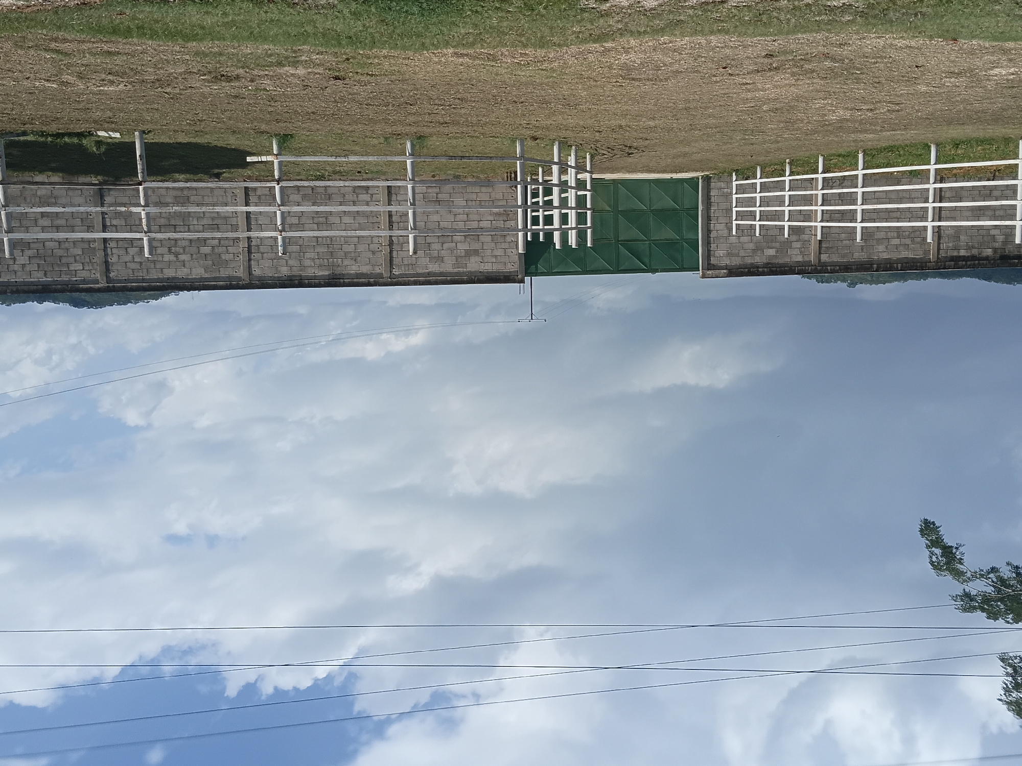 The image shows a horse paddock in Taguanes, Venezuela, with a brick wall in the background. A large green metal gate is visible in the center of the wall. White fences line the paddock, and the ground is covered in dry grass and dirt. The sky is cloudy, with some patches of blue. There are power lines strung across the top of the image. There are no people or animals visible in the image.