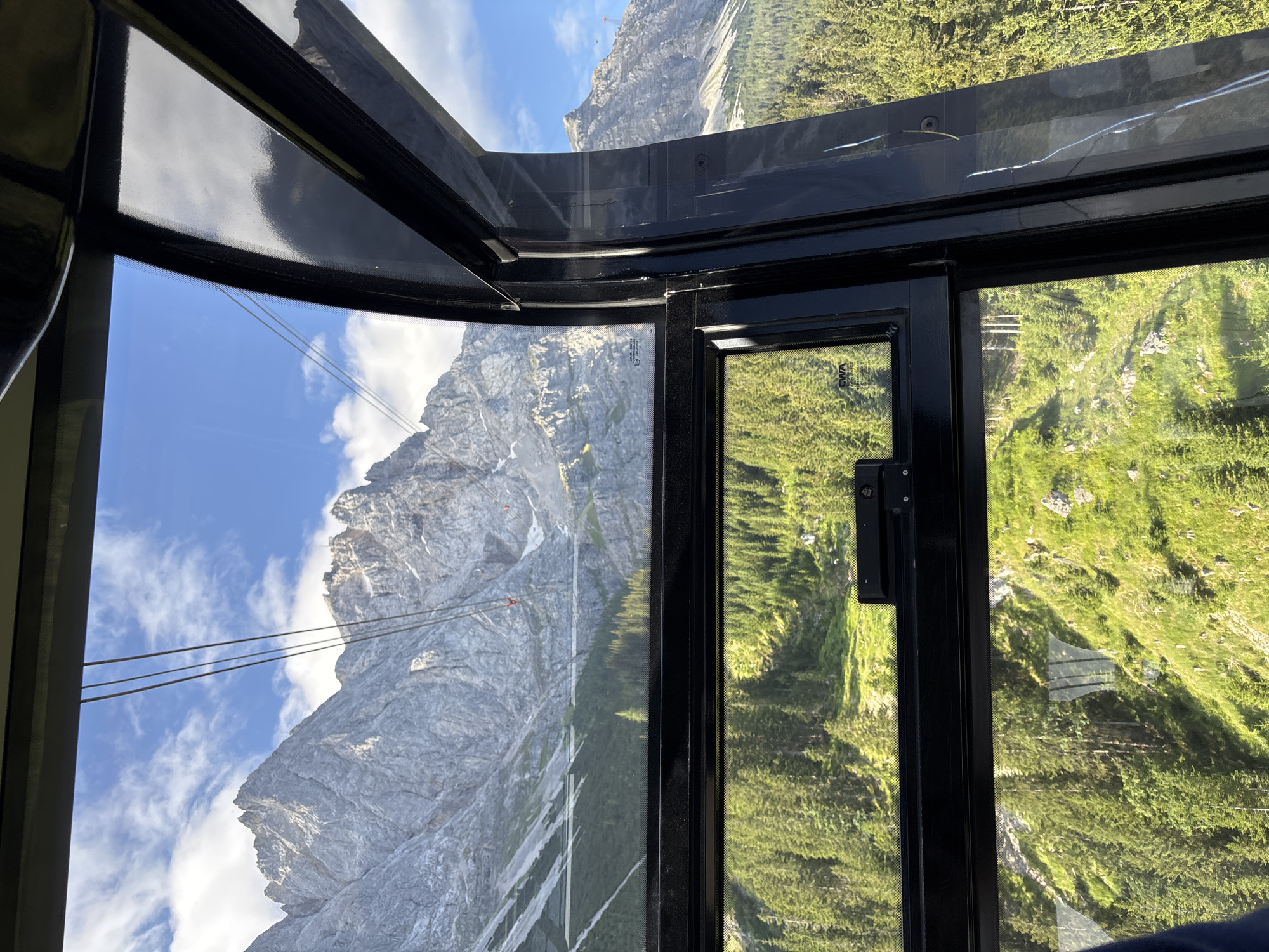 An image taken from inside a cable car shows a panoramic view of the Zugspitze mountain range near Grainau, Germany. The cable car's interior frame is visible, with black metal accents and large glass windows. Through the windows, the rugged, rocky peaks of the Zugspitze dominate the background, under a bright blue sky with scattered white clouds. The middle ground is covered with a dense forest of evergreen trees, which transitions into a lush green, grassy slope dotted with rocks. Multiple steel cables for the cable car system stretch across the scene, disappearing into the distance towards the mountains. In the upper reaches of the mountains, structures that appear to be buildings or observation decks are visible. The overall scene depicts a scenic mountain landscape viewed from a cable car.