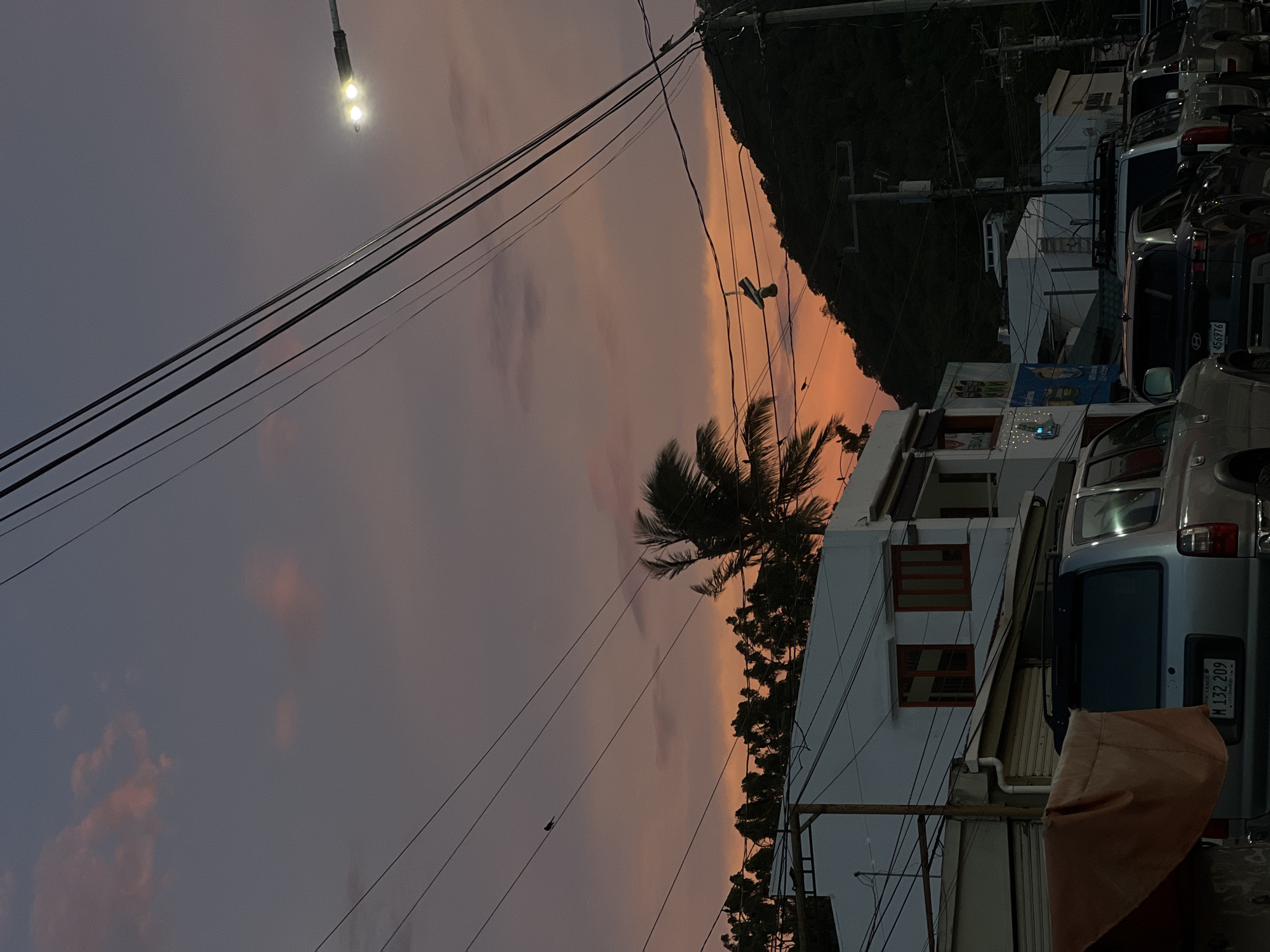 This image captures a street scene in San Juan del Sur, Nicaragua, during sunset. The sky displays a gradient of soft purples and oranges, with a few wispy clouds. Power lines crisscross the frame, a common sight in urban environments. A street lamp illuminates a portion of the sky on the right side.
In the foreground, several vehicles are parked along the street. The most prominent is a silver SUV with a license plate reading "M 132 209" and "NICARAGUA" below. To its right, other vehicles are partially visible.
Buildings line the street. On the left, a white building with orange-trimmed windows is visible. Adjacent to it is another building with a storefront, and further down, more structures can be seen. A palm tree with its fronds swaying in the breeze is positioned behind the white building.
A dark, tree-covered hill dominates the background on the right side of the image. The overall impression is a tranquil evening in a coastal town.