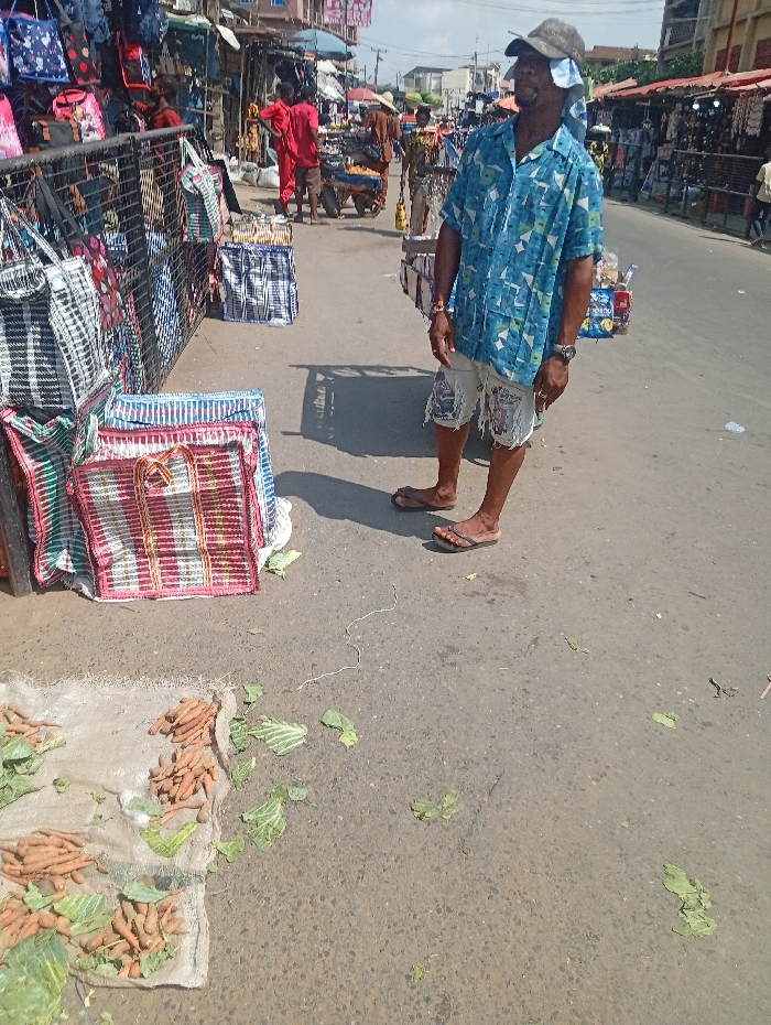 The image depicts a bustling open-air market street scene in Onitsha, Nigeria, under daylight conditions.
In the foreground, an adult male stands facing right, wearing a light blue patterned short-sleeved shirt, distressed light-colored shorts, a khaki baseball cap with a white cloth draped over his neck, and dark flip-flops. He has a dark wristwatch on his left wrist and a bracelet on his right. His expression is directed upwards and to the right. To his left, on the asphalt road surface, are piles of light brown root vegetables (possibly carrots or a similar tuber) and scattered green cabbage leaves, displayed on a piece of light-colored fabric or sack.
Immediately behind this produce, and to the left of the image frame, is a display of numerous woven plastic reusable shopping bags, stacked and hanging from a metal barrier or fence. These bags feature various stripe patterns, predominantly in black and white, and red and white with green and yellow accents.
The midground shows a street extending into the distance, lined with market stalls and additional people. Several individuals are visible; one in red attire is standing near a merchandise cart. Further down the street, more vendors and structures with awnings are present, suggesting a continuous commercial activity. The sky is bright and appears mostly clear. The overall environment is characterized by commercial activity, merchandise displays, and pedestrian traffic on an asphalt road.