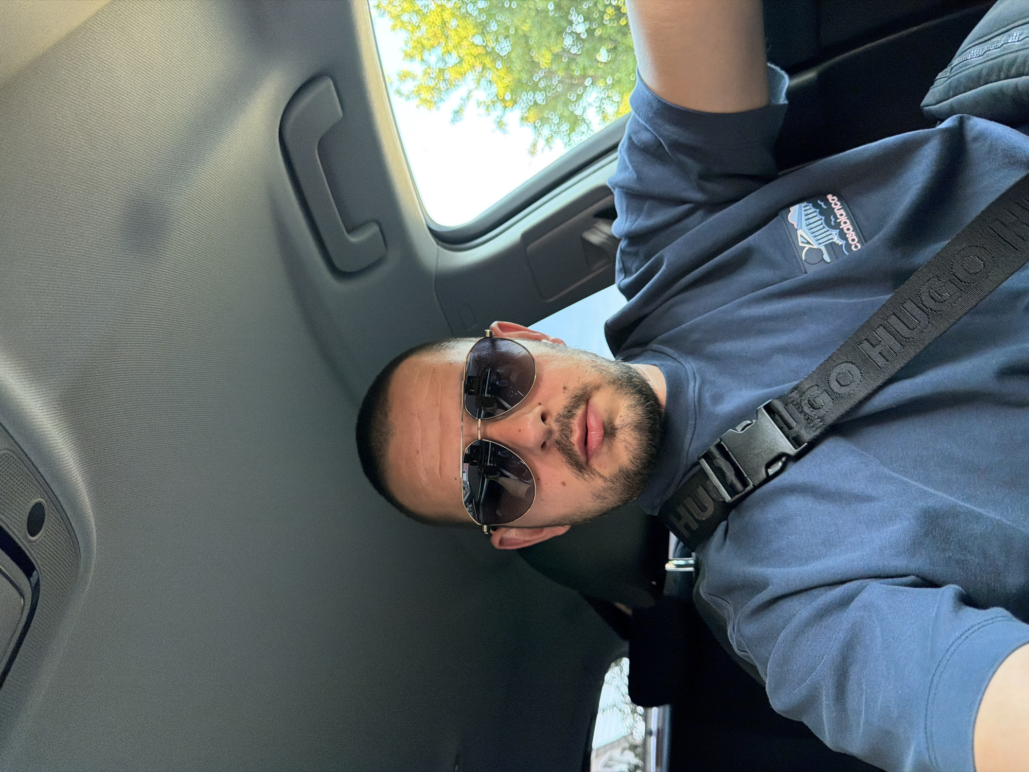 A man with a short haircut and a beard is sitting in the driver's seat of a car, wearing aviator sunglasses and a dark blue t-shirt with a "casablanca" logo on it. A black seatbelt with "HUGO" written on it is visible across his chest. The interior of the car, including the ceiling and a grab handle, is visible. Sunlight streams in from the right side of the car, illuminating the interior and the trees outside. The man appears to be looking at the camera. The image was taken in the city of Burgas, Bulgaria.