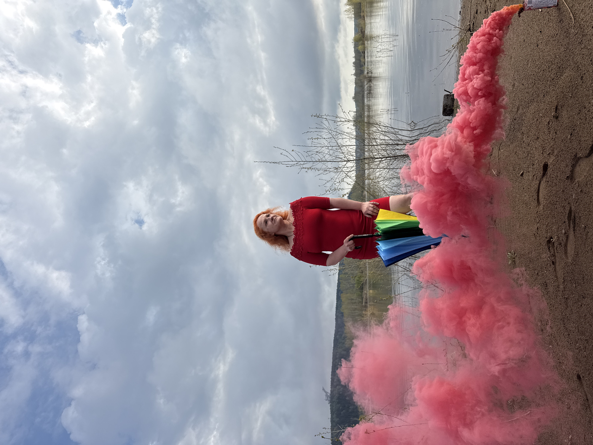 A red-haired woman in a red dress stands beside a body of water, likely a lake or river, in Noviy, Russia. She holds a multicolored umbrella and a pink smoke grenade. The grenade emits a large plume of pink smoke. The sky is cloudy. The ground is sandy. The water reflects the sky. Bare tree branches are visible behind the woman.