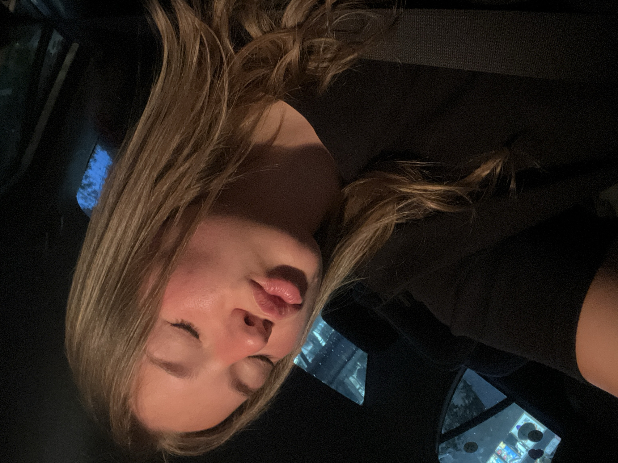 This close-up, vertically oriented image depicts a young woman resting within what appears to be a vehicle interior. Her long, wavy light brown or blonde hair frames her face, and her eyes are gently closed, suggesting she is asleep or in a peaceful state. Her lips are slightly parted, and her skin is fair. She is wearing a dark, possibly black, garment.
The background is predominantly dark, indicating low light conditions, likely nighttime. Visible through the windows to the left and right of her head are reflections of urban lights and structures, some with a distinct bluish glow. These reflections suggest the vehicle is in an urban environment. Dark, textured surfaces within the vehicle's interior are also visible. The overall mood is calm and serene. No discernible text is present in the image. This scene is set in Budva, Montenegro.