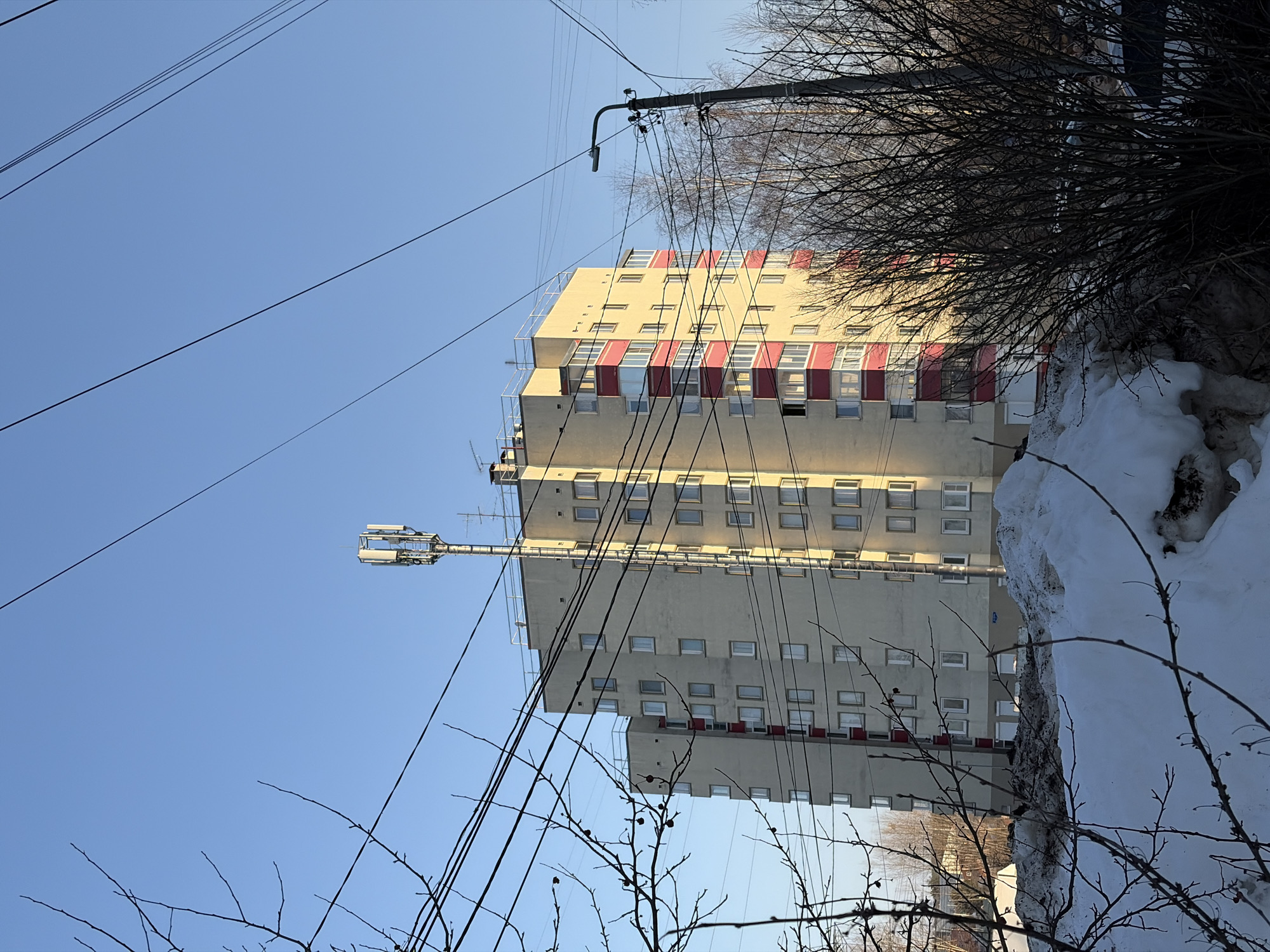 The image presents an outdoor urban scene in Chaikovsky, Russia, captured during what appears to be late winter or early spring. The primary subject is a multi-story residential building, approximately ten floors in height, constructed of light beige material. It features numerous rectangular windows and balconies with prominent red-framed railings. A cellular communications antenna array is affixed to the roof of this building. Direct sunlight illuminates the building from the right, creating contrasting shadows. In the immediate foreground, large, irregular piles of partially melted, soiled snow are visible. Numerous bare tree branches, some bearing small dark berries, extend into the frame from the left, partially obscuring the view of the sky and the building. Multiple black utility wires traverse the clear blue sky diagonally. To the right of the central building, a dark utility pole supports a street light and a dense network of attached wires. Additional bare trees and indistinct low-rise structures are discernible in the background. No individuals or specific activities are depicted within the scene.