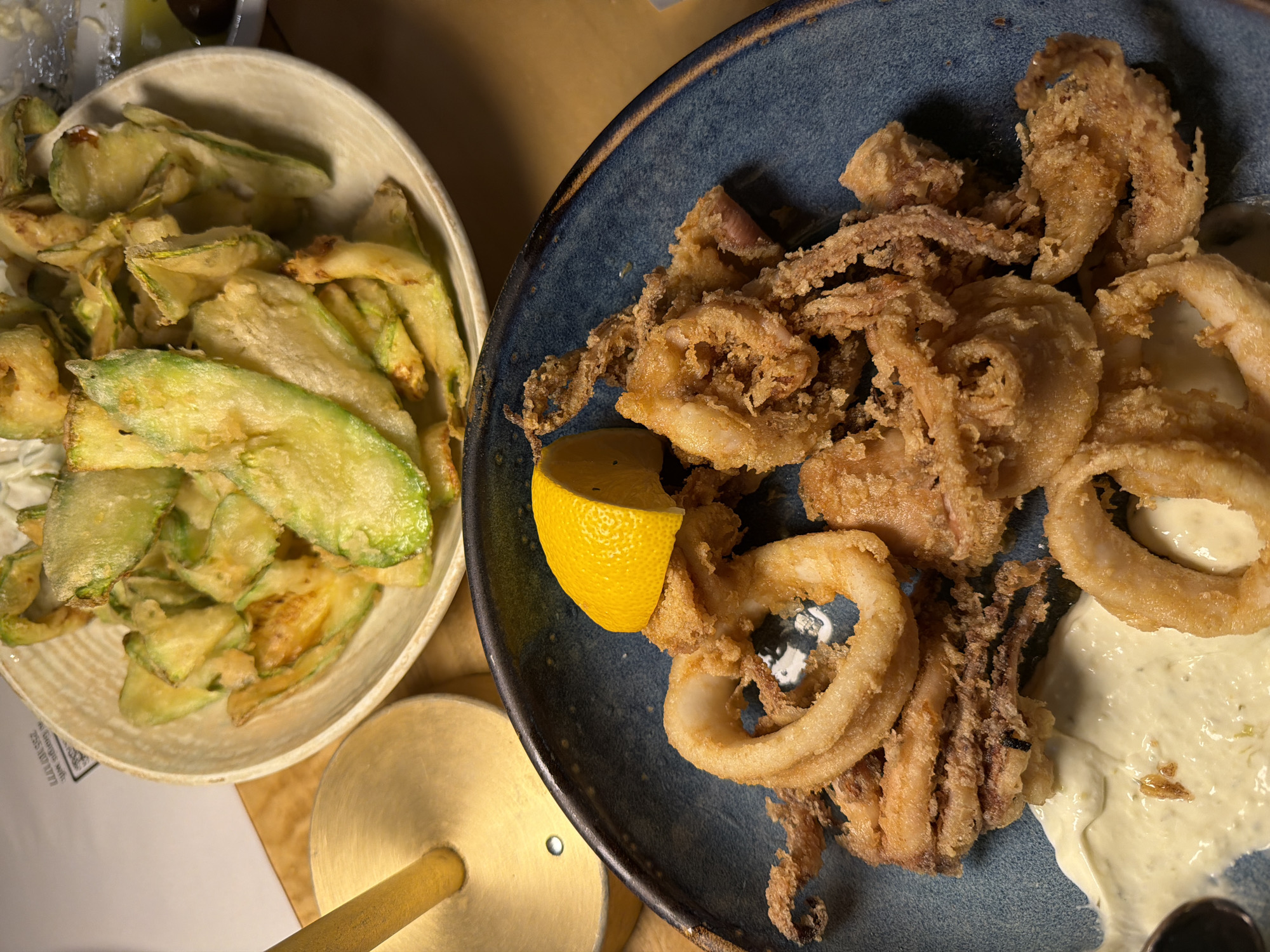 An overhead shot captures a meal laid out on a table, likely in a restaurant setting in Makri, Greece. The primary subjects are two dishes: a bowl of fried zucchini slices and a plate of fried calamari.
The left dish features a light-colored, oval-shaped bowl filled with numerous thin, crispy-looking slices of fried zucchini, which have a light green and golden-brown hue. To the right, a dark blue or grey, textured plate holds a generous serving of fried calamari, consisting of both rings and tentacles, all coated in a light, crispy batter. A bright yellow lemon wedge is nestled among the calamari, and a dollop of creamy white sauce, possibly aioli or tartar sauce, is visible on the plate, partially beneath some of the calamari pieces.
The table surface appears to be wooden, and the lighting is warm and artificial, suggesting an evening meal or an indoor ambiance. Part of a white paper or receipt is visible beneath the zucchini bowl, showing fragmented text that includes "georgia md." and a series of numbers starting with "235 000 ????". A golden or brass-colored object, possibly the base of a lamp or stand, is seen in the lower left corner of the frame. The scene presents a classic Mediterranean appetizer spread.