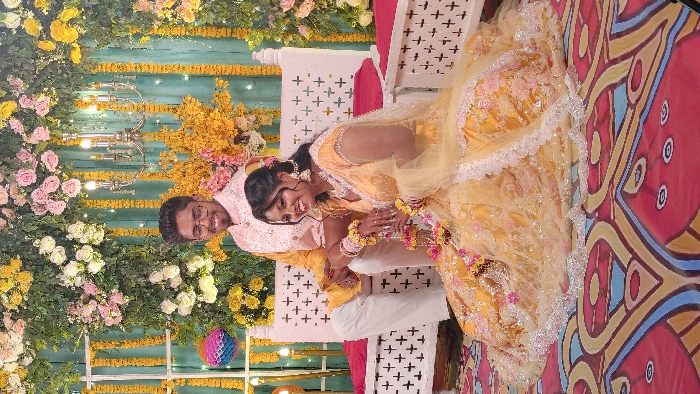 An indoor event in Dhaka, Bangladesh, features a young adult couple against a decorative floral backdrop. The male, approximately in his late 20s, is seated on a white carved wooden bench. He wears eyeglasses, a light pink embroidered jacket over a yellow kurta, and white trousers, with a yellow floral wristband on his right wrist. He maintains a direct gaze and a slight smile towards the camera.
The female, also in her late 20s, is seated on the floor directly in front of and to the right of the male. She wears an elaborate yellow traditional outfit with extensive embroidery, a matching sheer dupatta, and significant traditional jewelry including large earrings, a necklace, bangles, and a yellow floral wristband on her left wrist. She holds a small, multi-colored floral ornament with tassels in her hands. Her head is turned slightly to her left, and she is smiling.
The backdrop consists of a dense arrangement of green foliage, white, pink, and yellow flowers, with vertical yellow floral garlands. A silver-toned chandelier with multiple electric candles hangs centrally behind the male. The white wooden bench features carved cross-shaped patterns and red fabric cushions. The floor is covered by a vibrantly patterned carpet in red, blue, yellow, and white geometric designs. A multi-colored paper lantern is visible to the left of the male.