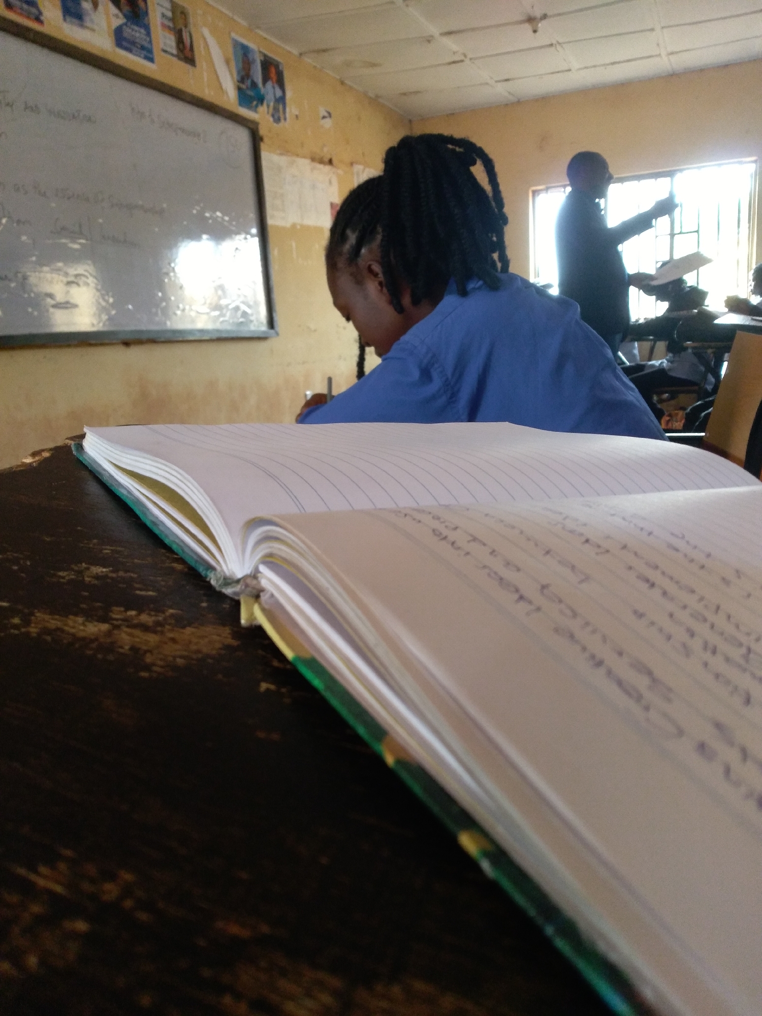 In a classroom in Jos, Nigeria, an overhead view shows an open notebook on a dark, textured wooden desk. The notebook's pages are lined, and the right-hand page contains handwritten text in purple ink. In the background, a person with dark braided hair, wearing a blue shirt, is seated. Further back, a teacher stands by a window, holding a document and gesturing. A whiteboard with writing is visible on the left wall, and framed pictures are posted above it. The ceiling is made of acoustic tiles, and a fluorescent light fixture is mounted to it. The overall scene depicts a typical classroom environment.