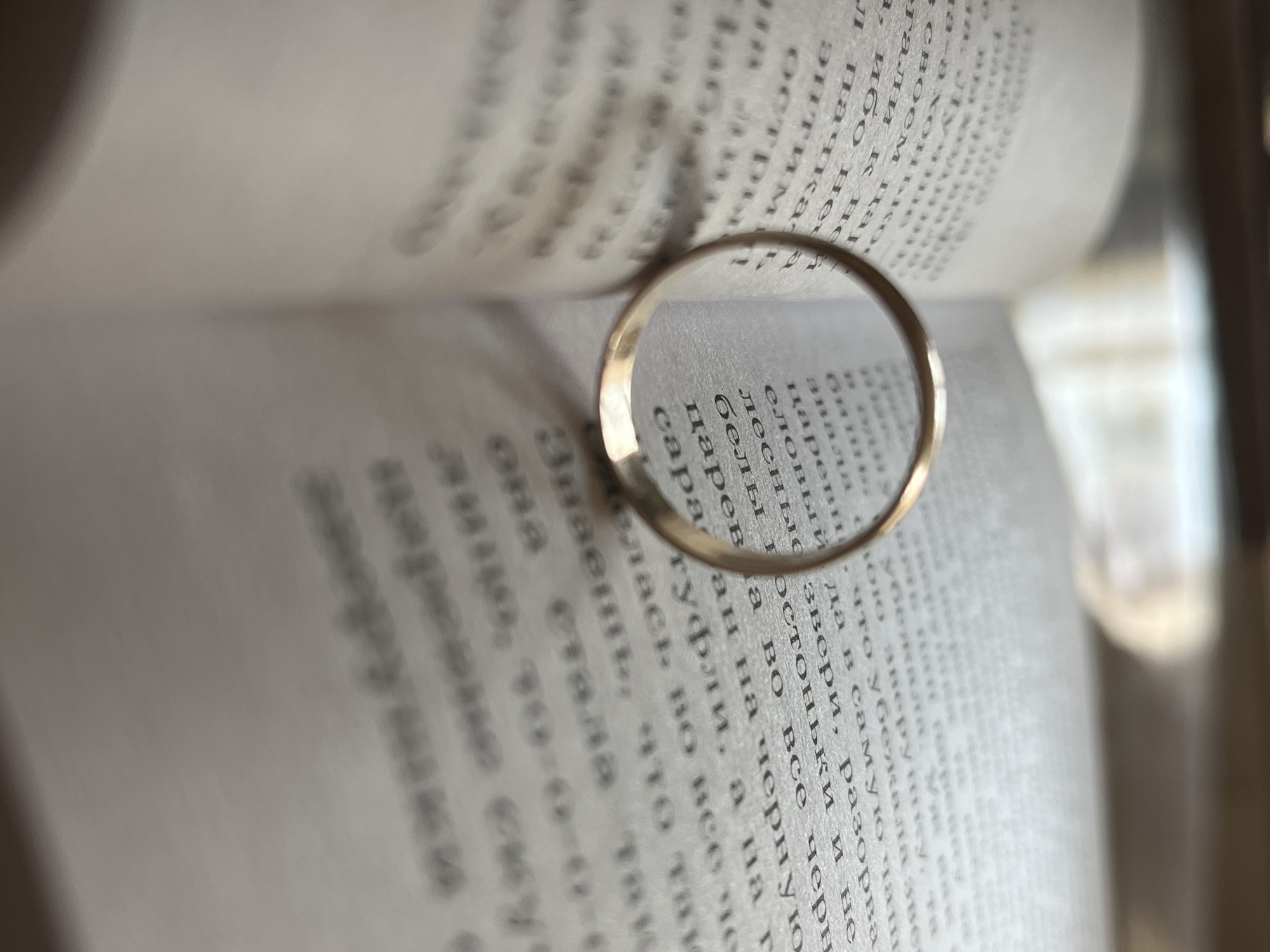 An extreme close-up shot focuses on an open book with a gold ring placed on its pages. The book is open to reveal text in a foreign language, with the text on the left page slightly out of focus and the text on the right page in sharp focus, framing the ring. The ring itself is a simple, unadorned band that catches the light, highlighting its metallic surface. The background is blurred and out of focus, suggesting a shallow depth of field and drawing all attention to the book and ring. No specific location within Krasnyye Barrikady, Russia, can be identified from the image.