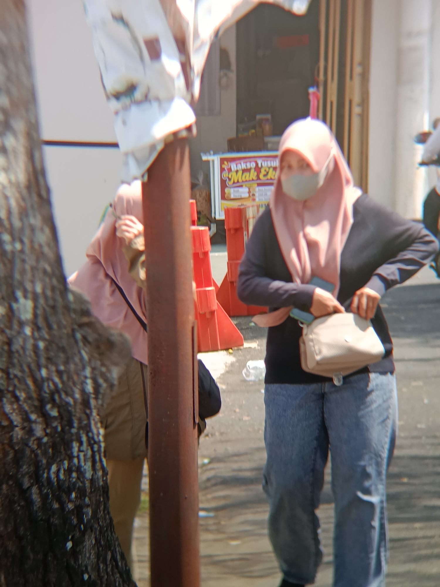 In Desa Kebon Manis Kecamatan Cilacap Utara, Indonesia, a person wearing a pink hijab and a gray face mask is seen from the waist down. They are wearing a dark long-sleeved shirt, blue denim jeans, and a beige crossbody bag. The individual is standing on a paved surface, with a red traffic barrier visible behind them. A tree trunk is in the foreground on the left, and a metal pole stands between the person and the barrier. In the background, a food stall with a sign that reads "Bakso Tusuk Mak Eki" is partially visible. The lighting suggests it is daytime.