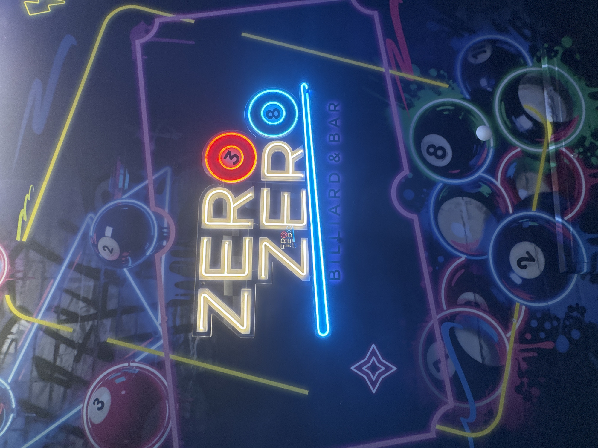 A nighttime view of a neon sign displaying "ZERO ZERO BILLIARD & BAR" in Varna, Bulgaria. The word "ZERO" is vertically stacked twice, with the top zero being represented by the number 3 and the second by an 8 ball. The background includes images of billiard balls and neon lights, suggesting a billiard or pool hall.