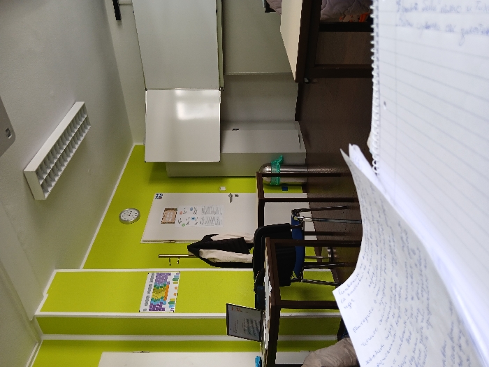 The image depicts the interior of a vacant classroom or study room. The specific location within Kaiserslautern, Germany, is not discernible from the visual evidence.
The room features bright lime green walls with white trim, a white ceiling with a centrally mounted rectangular fluorescent light fixture, and a dark brown floor. In the immediate foreground, two open spiral-bound notebooks with handwritten text in a Latin script are positioned on a desk. To the left-midground, a dark brown rectangular table supports an open silver-colored laptop and a dark object, with a dark blue chair pulled beneath it.
On the left wall, a white door with a silver handle is visible, adorned with several printed papers. Above and to the right of this door, a round analog clock with a white face and black numerals is mounted. A metallic coat rack with a black jacket and a light-colored garment hangs near the door. Further back in the room, two large, blank whiteboards are mounted on the far wall. A silver cylindrical trash can with a green plastic bag liner is situated on the floor near the far wall. A colorful chart or poster is affixed to the lime green wall section to the left of the door. No individuals are present in the scene.
