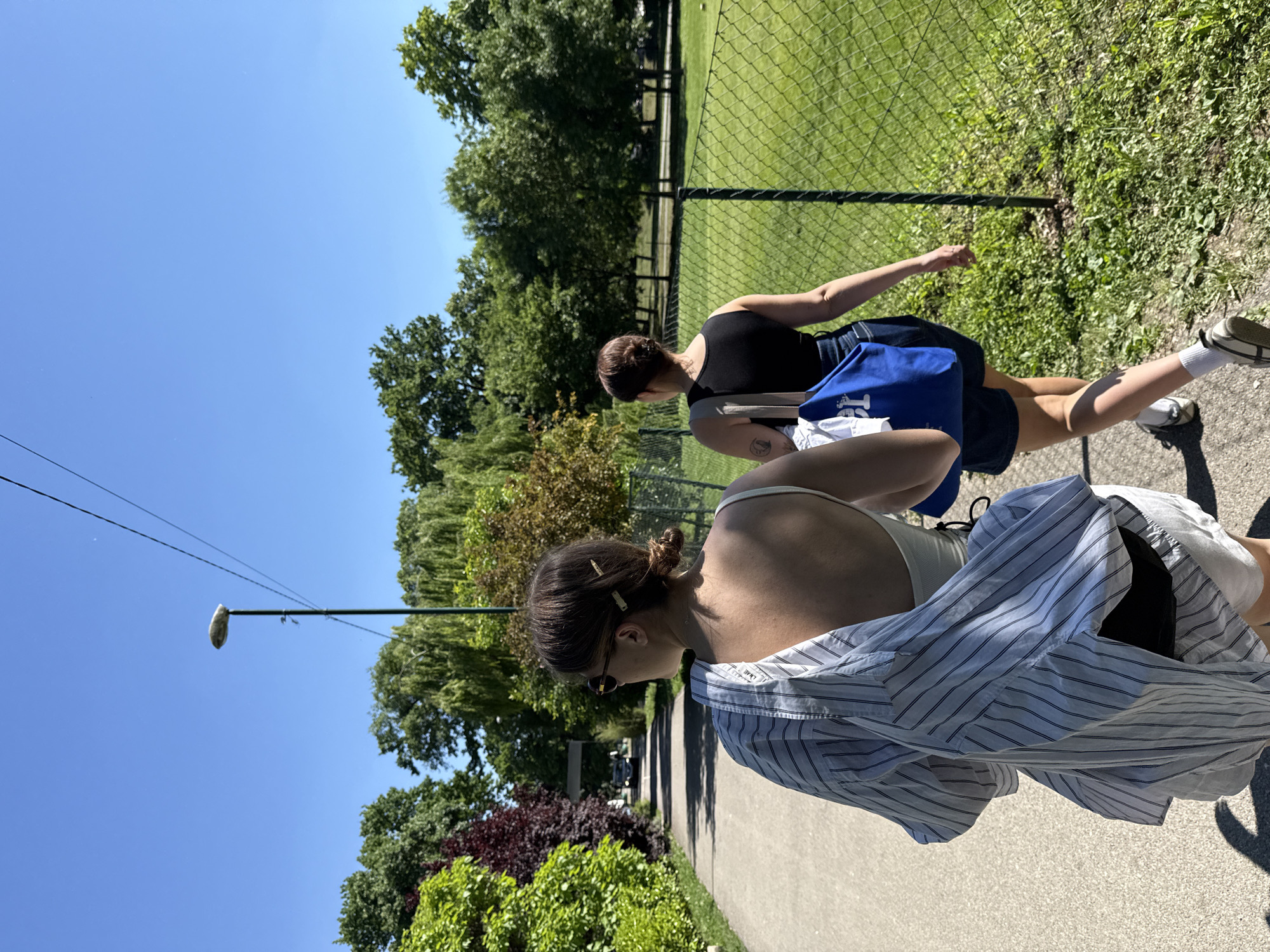 The image shows two women walking on a sidewalk in Cukarica Urban Municipality, Serbia. The first woman, closer to the viewer, wears sunglasses, a white tank top, and a blue and white striped shirt. The second woman wears a black tank top and a dark skirt, carrying a blue bag. A chain-link fence separates the sidewalk from a grassy area with trees in the background. A street lamp is visible on the left side of the image. The sky is clear and blue.