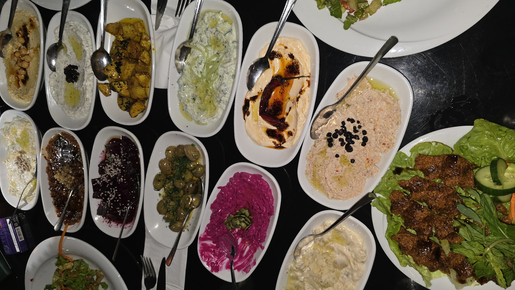 The image presents an overhead view of a diverse array of small dishes, commonly known as mezzes or appetizers, meticulously arranged on a dark, possibly black, table. There are at least 15 different dishes, primarily served in white oval or rectangular bowls and plates, creating a vibrant culinary spread. Among the visible dishes, starting from the top left, is a bowl of hummus garnished with whole chickpeas and a dark drizzle. Next to it, a creamy white dip, possibly a yogurt-based spread like haydari, is adorned with black specks (perhaps olives or nigella seeds) and olive oil. Further right, golden-brown roasted potatoes are sprinkled with black sesame seeds. Continuing right, a light green cucumber and yogurt dip, akin to cacık or tzatziki, is topped with fresh cucumber strands and olive oil. In the center of the frame, a smooth, light orange dip, possibly a spiced hummus or a variant of muhammara, is richly garnished with a dark red, spiced oil. Below this, a pinkish-red dip, perhaps a spicy pepper paste, is decorated with small black currants or capers and olive oil. To its right, a large white plate holds several servings of çiğ köfte (spiced bulgur patties, often served raw) nestled on fresh lettuce leaves, accompanied by cucumber slices and green herbs. On the bottom row, a striking bright magenta-colored dip, likely a beetroot spread, is topped with green pumpkin seeds. Next to it, vibrant green olives are marinated with fresh dill. Further left, a dark red beetroot salad is garnished with white sesame seeds. A glossy, dark brown dish, possibly a lentil pâté or another nut-based spread, is topped with crushed nuts. Another creamy white dip, likely a walnut or fava bean spread, is garnished with chopped walnuts and olive oil. At the very bottom right, a similar white dip is topped with almond slivers. Two partial plates of fresh green salad are visible at the top right and bottom left, suggesting a larger meal. Most dishes include spoons, indicating they are meant for communal sharing. The overall setting appears to be an indoor dining environment, possibly a restaurant or a festive home meal, given the extensive variety and presentation. The lighting is warm, enhancing the colors of the food. A white napkin and some cutlery (a fork and knife) are visible on the right side. A small, partially obscured purple card with "TER..." visible text is seen in