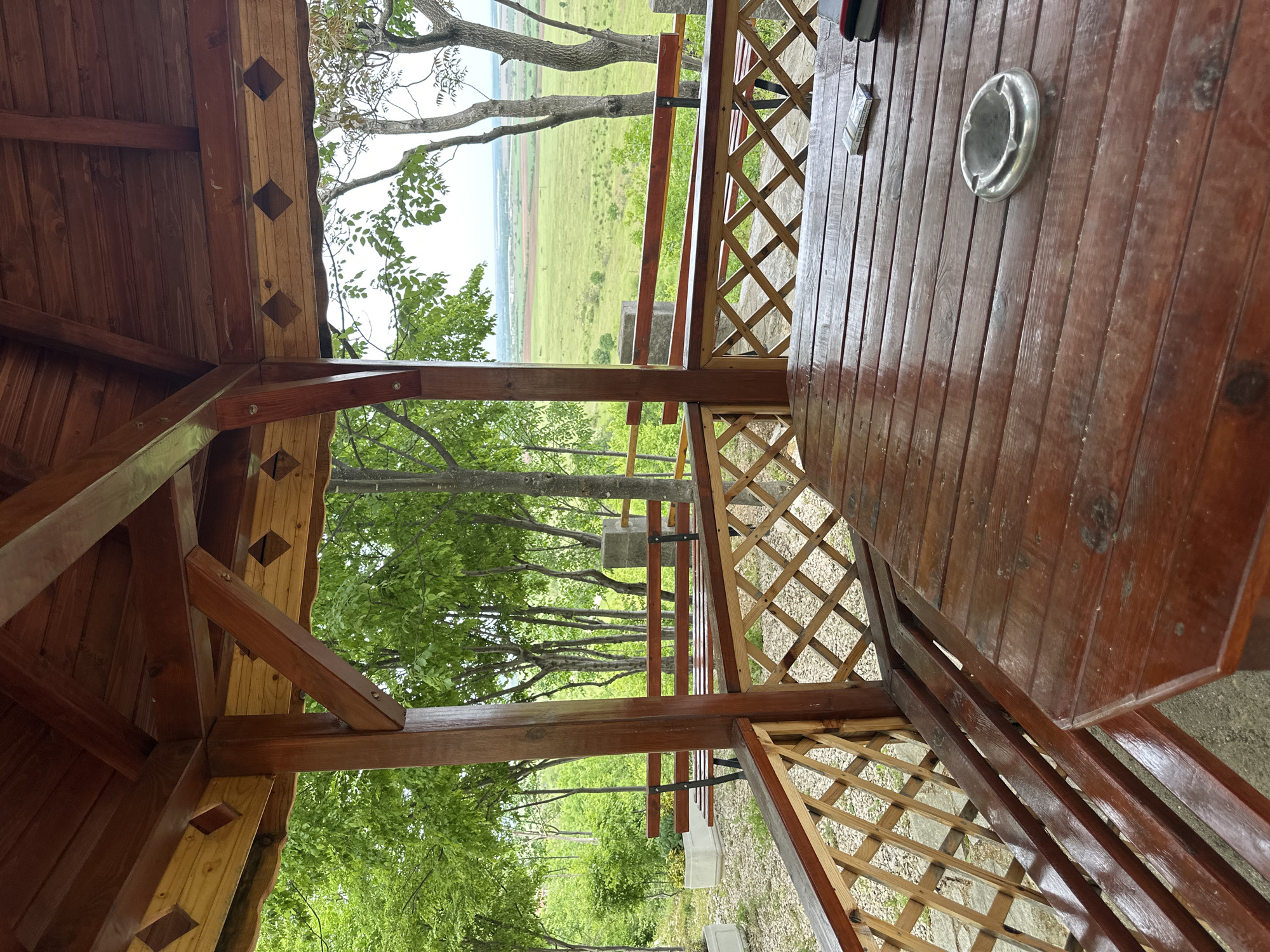 The photograph depicts the interior of a wooden gazebo located in the city of Gara Oreshets, Bulgaria. The structure features a lattice railing and a wooden table, upon which sits a silver ashtray and a packet of cigarettes. Outside the gazebo, a landscape of green fields and trees is visible. The gazebo structure includes wooden beams with decorative diamond cutouts.