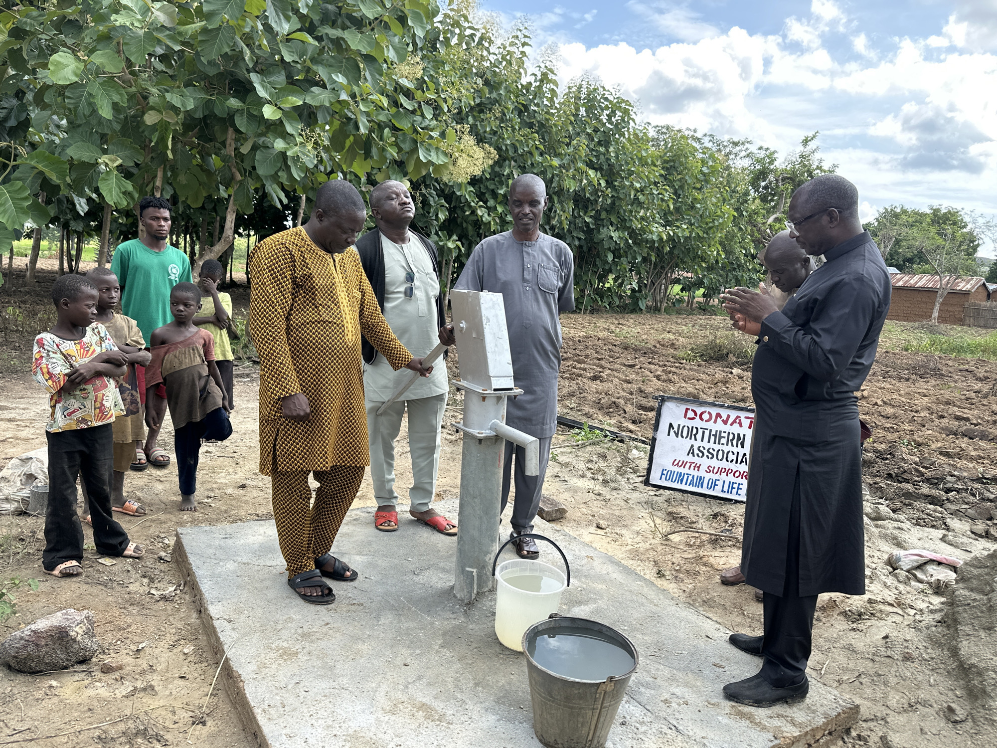 In the city of Afogo, Nigeria, a group of individuals, including several young children and adult men, are gathered around a newly installed water pump. The scene appears to be a communal area, possibly a village square or a designated spot for the water source, with a dirt ground and a few buildings in the background. The sky is partly cloudy.
The water pump itself is a hand-operated model, made of metal, mounted on a concrete base. Two buckets are placed on the base, one white and a bit smaller, the other a dark metal bucket. The white bucket contains clear water, while the metal bucket holds a larger quantity of water.
The people present are dressed in a mix of traditional and modern attire. A man in a bright yellow and black patterned robe stands closest to the pump, facing away from the camera. To his left, a man in a grey outfit with a similar buttoned collar is looking down, perhaps in prayer or contemplation. Further left, two young boys and a girl are looking towards the pump. Another man in a green t-shirt is standing behind them, observing.
To the right of the pump, a man in a black outfit, possibly a traditional robe with a split skirt, stands with his hands clasped, facing the pump. Another man, partially visible behind him, also appears to be dressed in dark clothing.
A white sign with black lettering is propped up on the ground behind the group on the right. The visible text reads "DONAT NORTHERN ASSOCIA WITH SUPPOR FOUNTAIN OF LIFE". This suggests the water pump may be a recent donation to the community.
The environment around the pump is characterized by dry, tilled earth, indicating it could be a farming area. Lush green trees and bushes are present in the background, providing a natural backdrop to the scene. The overall impression is one of a community gathering to inaugurate or inspect a new, vital resource.