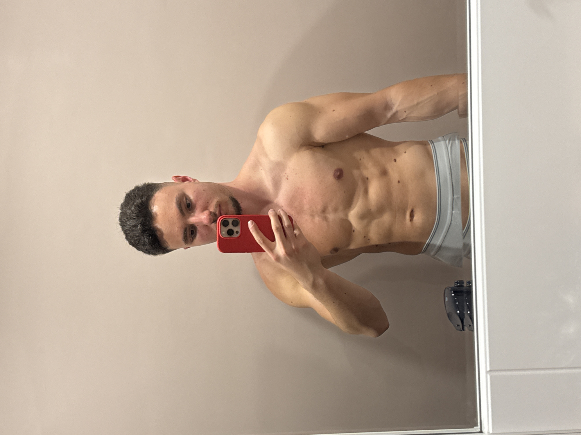 A man is captured in a mirror selfie, shirtless and displaying a muscular physique, including defined abdominal muscles and visible veins in his arms. He has short, dark hair and stubble/beard. He is wearing light-colored boxer briefs with horizontal stripes. The man holds a red smartphone, possibly an iPhone given the multi-lens camera array, with his right hand, pointing it towards the mirror to take the photo. His gaze is directed towards the phone screen, implying he is looking at his reflection.
The setting appears to be an indoor room with plain, light-colored walls, possibly a bathroom or bedroom, as indicated by the mirror and a white cabinet or door frame on the right side of the frame. A dark-colored object, possibly a small bottle or container, is partially visible near the bottom right edge, resting on a surface next to the mirror. The lighting in the room is soft and even. No specific time of day or weather conditions can be determined from the image. There is no visible text. The man's expression is neutral. The original image is rotated 90 degrees counter-clockwise. The location context is Sofia, Bulgaria.