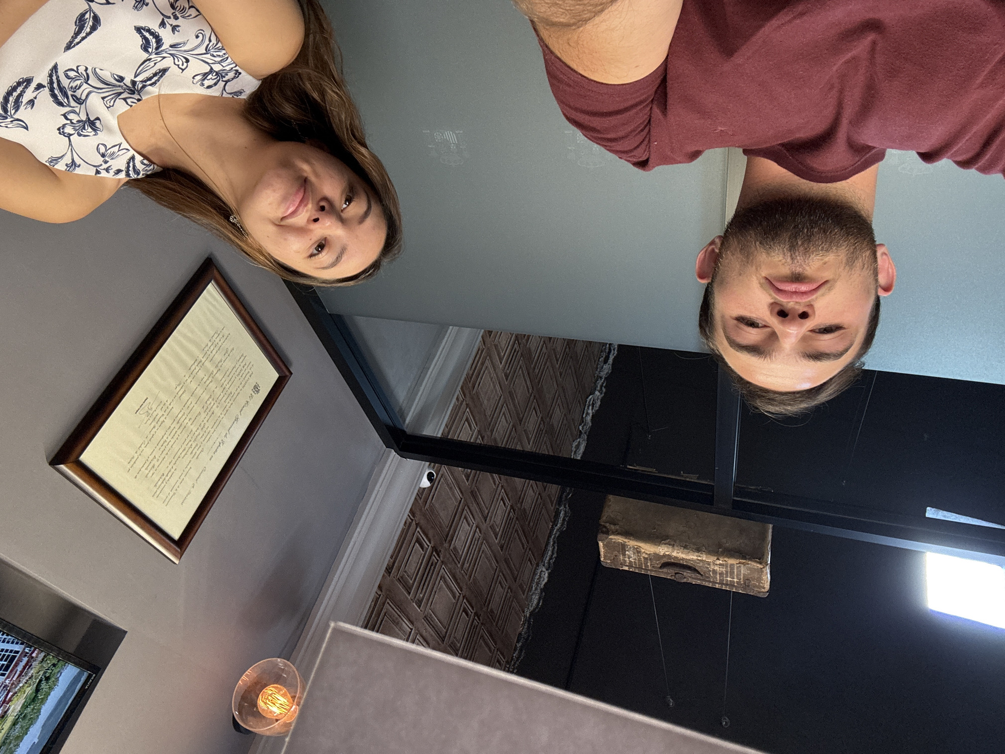 This image presents a unique, low-angle perspective of two people, a woman and a man, who appear upside down, looking down towards the camera. The woman, on the left, wears a white sleeveless top with a dark blue floral pattern and has a soft smile. The man, on the right, has a beard, wears a maroon t-shirt, and holds a neutral expression.
The setting is an indoor, modern space, likely a commercial or public establishment in Apetatitlán, Mexico. The background features grey walls and ceiling. Several distinct objects are visible:
- On the left wall, a framed document with legible text in Spanish, starting with "En el Gran Salón General de la Legión..."
- Partially visible below the document is a wall-mounted television displaying a landscape scene.
- A wall-mounted light fixture with an exposed Edison-style filament bulb.
- A small, white security camera is mounted high on a structural beam or wall.
- A textured surface with a brown diamond pattern, possibly a decorative wall panel or a reflection.
- A rustic, wooden, rectangular object, resembling an old suitcase or chest, hangs from the ceiling.
- A bright, rectangular light panel is visible towards the bottom right of the frame.
Faint geometric patterns are also reflected on the grey ceiling surface. The calm expressions of the individuals and the contemporary indoor decor, combined with the security camera and the formal Spanish document, suggest an event or casual gathering in a well-appointed, possibly historical or official, public space.