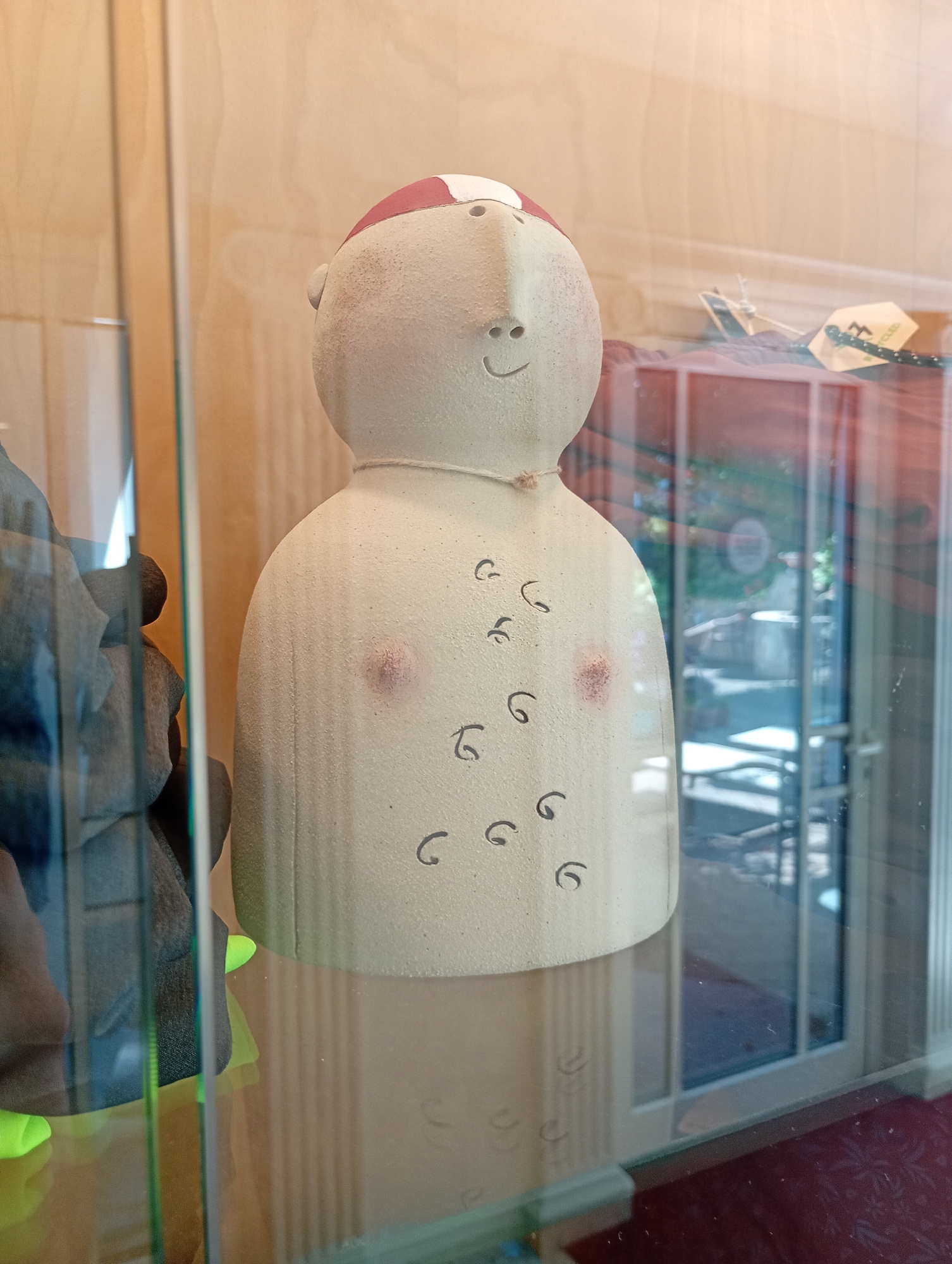 An unpainted ceramic figurine of a torso with a head is displayed in a glass case. The figurine has a red band across its head and simple facial features. Black, comma-like marks are scattered across the torso, with two reddish circles on the chest area, suggesting nipples. A thin rope or string is tied around the neck area. The figurine is positioned in the foreground, viewed through glass. In the background, through the glass, a wooden paneled wall and a door with glass panels are visible, suggesting an indoor setting like a shop or display area. The lighting is neutral. The overall environment appears to be a clean, indoor retail or exhibition space, possibly in Bad Sachsa, Germany, given the context of the query. There are no people actively interacting with the figurine, but reflections in the glass suggest the presence of a viewer or photographer.