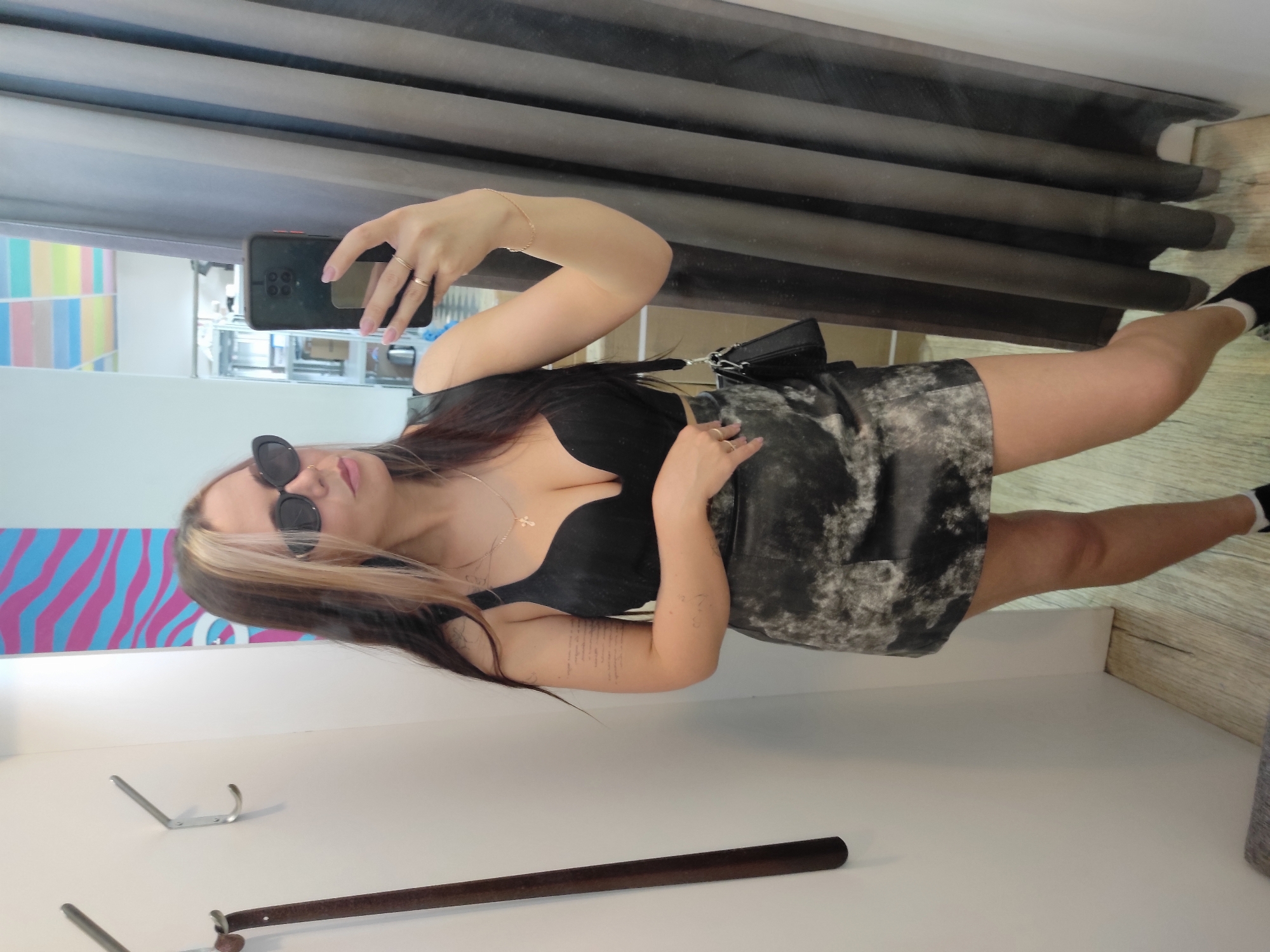 A young woman in a black top and grey patterned skirt is taking a selfie in what appears to be a fitting room. She is wearing sunglasses and a gold necklace. She has light and dark hair. There is a handbag over her shoulder. The background includes a blue and pink striped wall and grey curtain. A wooden rod with a metal hook lies on the white counter below. The location is Chaikovsky, Russia.