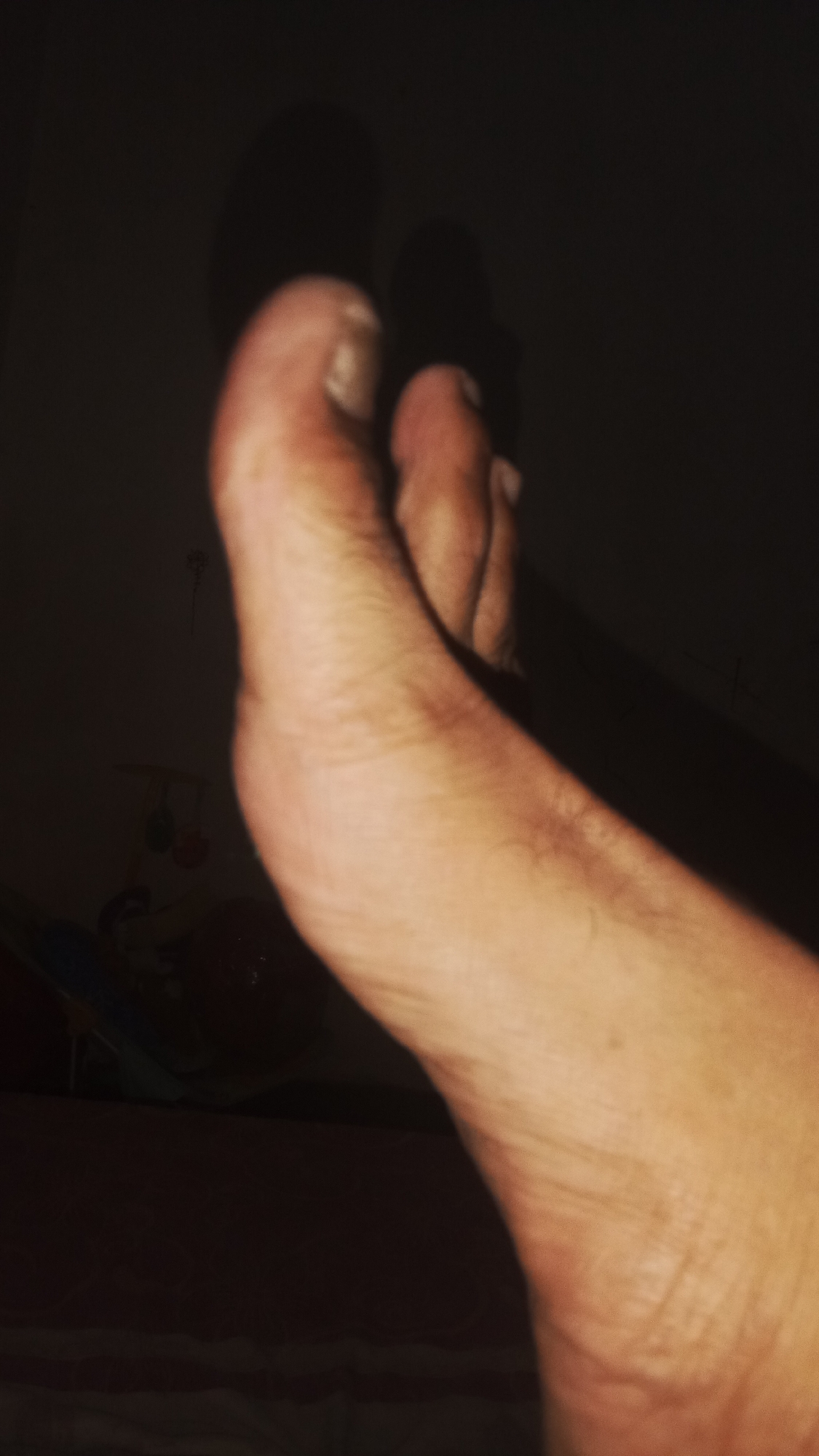 The image displays a close-up, dimly lit view of a human foot, likely positioned in an indoor setting, possibly within a bedroom. The foot is angled, with the toes prominently featured. The skin appears light-skinned with visible texture and some superficial lines. The background is dark and indistinct, with shadows obscuring details of the surroundings. A faint shadow of a mobile or hanging toy can be observed in the upper left portion of the image, suggesting the presence of a child's room or area. The overall lighting is low, creating a somber and intimate atmosphere. The location of Sungai Sipai, Indonesia, is not discernible from the visual information provided in the image.