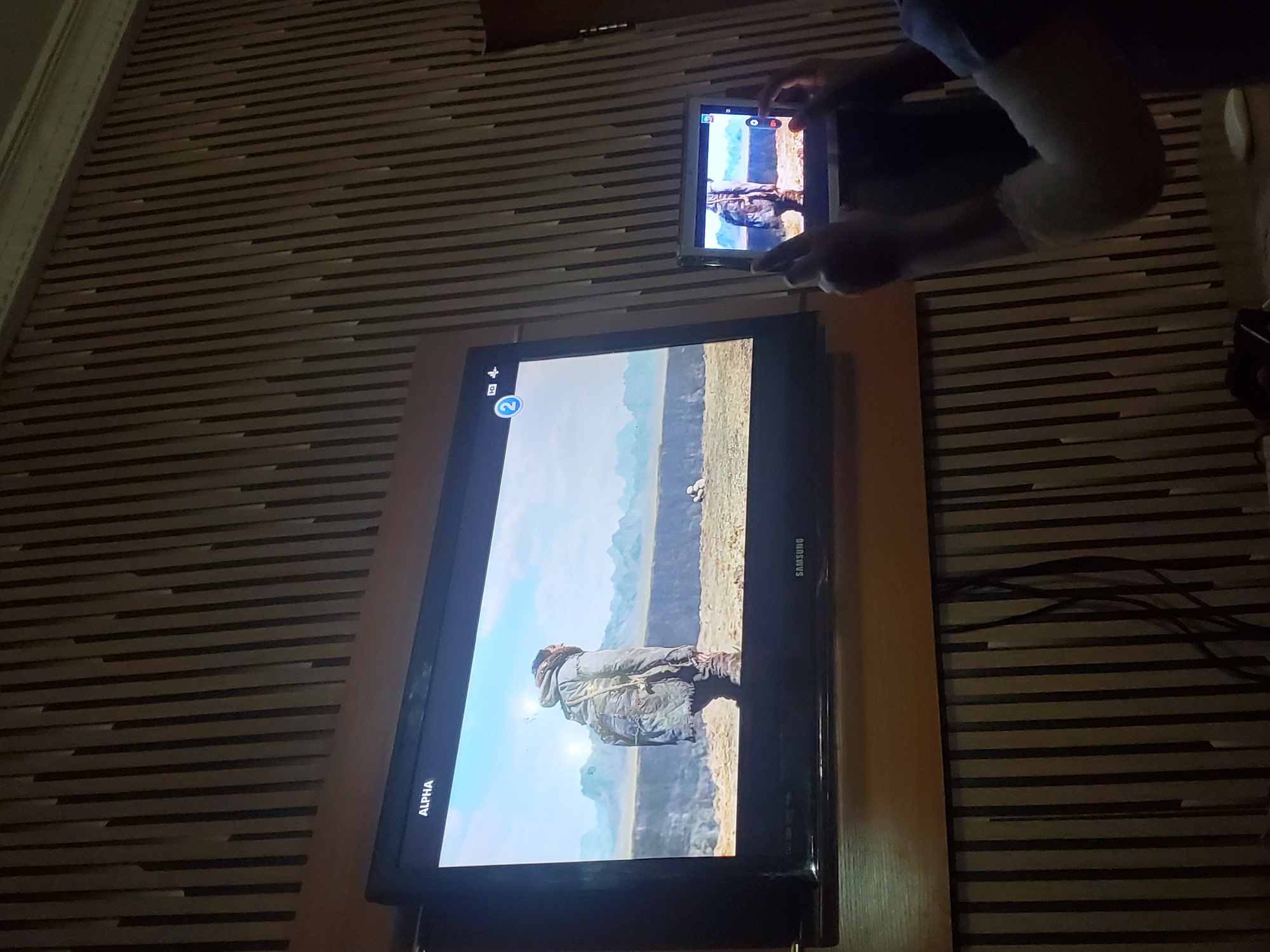 A person holds a tablet in front of a television screen displaying similar imagery. The tablet and television screen both show an individual in an outdoor setting. The environment appears to be a room with horizontally striped wallpaper. "Alpha" and "Samsung" are visible labels on the television. The location is unspecified, though it is noted to be within Obosi, Nigeria. The specific actions are viewing media on the tablet and television.