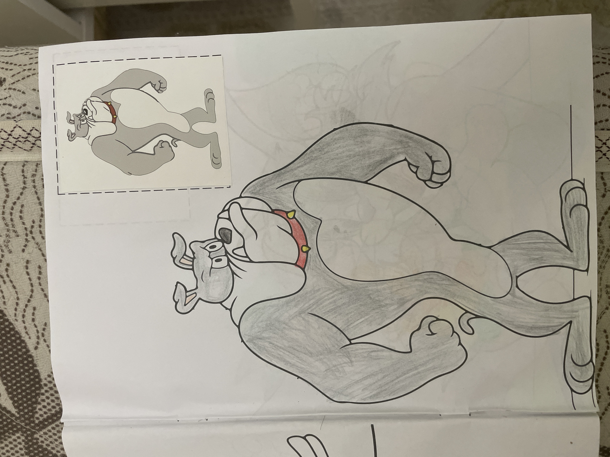 The media file displays a coloring book page, featuring a cartoon bulldog character resembling Spike from Tom and Jerry. The page contains two main depictions of the bulldog: a smaller, fully colored reference image in the upper left corner, enclosed by a dashed border, and a larger, partially colored version dominating the center-right of the page.
The reference image shows the bulldog with a gray body, white face and belly, a red spiked collar with yellow studs, and black outlines. The larger drawing is in the process of being colored; its body is shaded with gray crayon or pencil, and the collar is colored red with yellow studs, consistent with the reference. However, areas such as the bulldog's face, ears, and belly remain white, indicating incomplete coloring. Faint, uncolored pencil outlines of another cartoon character (possibly a cat) are visible in the background behind the larger bulldog, suggesting a multi-character scene yet to be completed.
The page itself appears to be resting on a textured, patterned surface, possibly a blanket or rug, indicating an indoor setting where a coloring activity is taking place. There is no visible text on the page. The image is oriented such that the characters are running upwards or sideways.