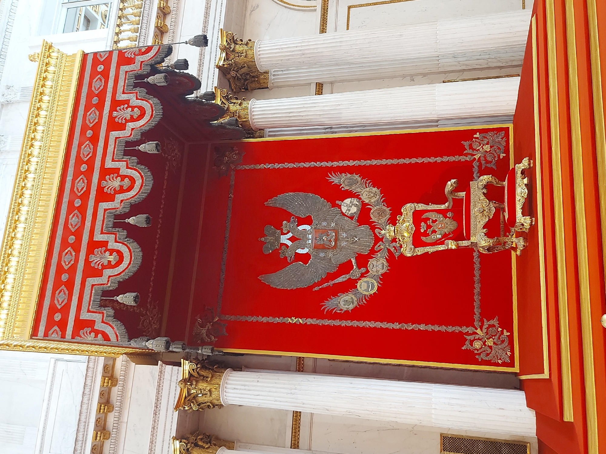 The image shows an ornate interior space with a red ceiling. The ceiling features a large, silver-colored double-headed eagle emblem in the center, along with other decorative silver and gold embellishments. The ceiling is bordered by gold trim. Supporting the ceiling are white columns with golden, intricately decorated capitals. The left side of the image shows a portion of the ceiling with a red and silver patterned design, bordered by gold trim. A red and gold curtain hangs on the right side of the image. The overall impression is one of opulence and grandeur. The location is unknown.