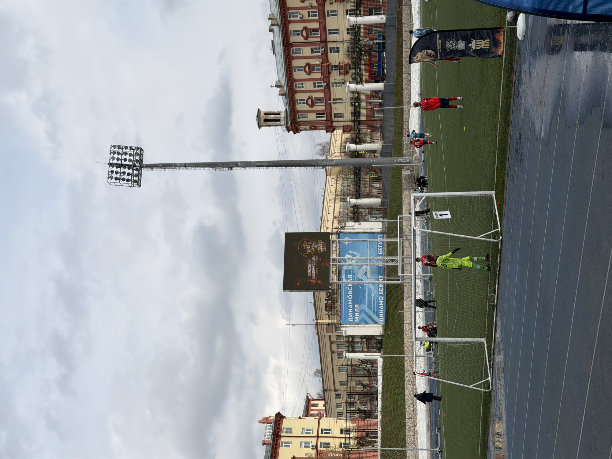 The image depicts a soccer field in Izhevsk, Russia, likely at the Dynamo stadium. A match or practice is in progress, indicated by players in red and other colored kits on the green field. A goalkeeper in bright green stands in front of a goal. Along the side of the field is a running track, and behind the field, there are buildings typical of Russian urban architecture. A tall lighting structure is visible, along with electronic scoreboards displaying information in Cyrillic script. The sky is overcast, suggesting a potentially rainy or cloudy day.