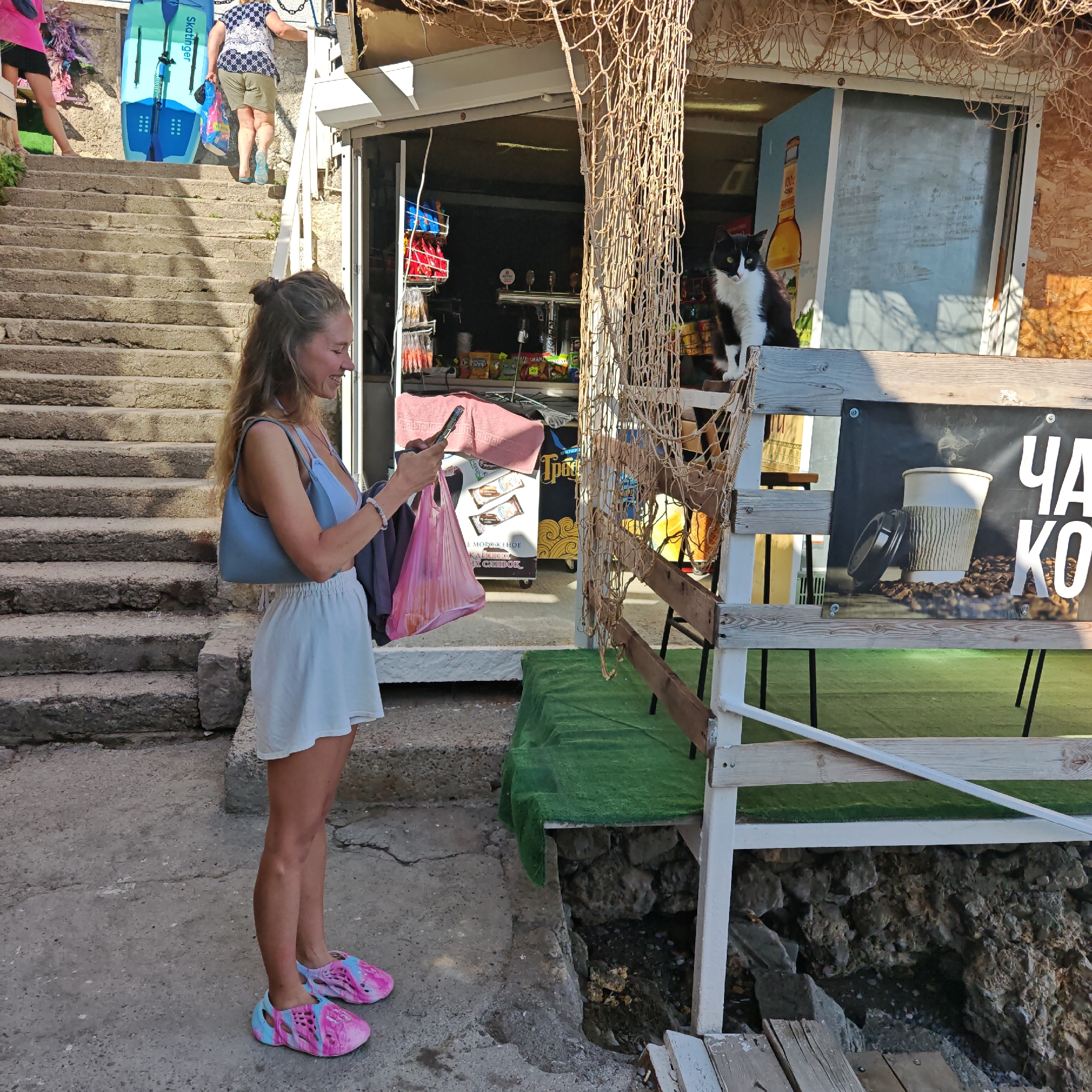 In the city of Simeiz, Ukraine, on a sunny day, a young woman with long blonde hair, wearing a light blue top and a white skirt, stands looking at her phone while holding two shopping bags, one pink and one dark gray. She is wearing multi-colored pink and blue crocs. Behind her, a black and white cat sits on a wooden ledge in front of a small shop. The shop is filled with various goods, including snacks and drinks. A net hangs in front of the shop, and a large sign displaying coffee and the words "CHA KO" is visible. The scene appears to be on a street with stone steps leading up to the shop, and a grassy area with wooden seating is set up in front of it. The overall impression is a lively, casual outdoor marketplace or cafe.