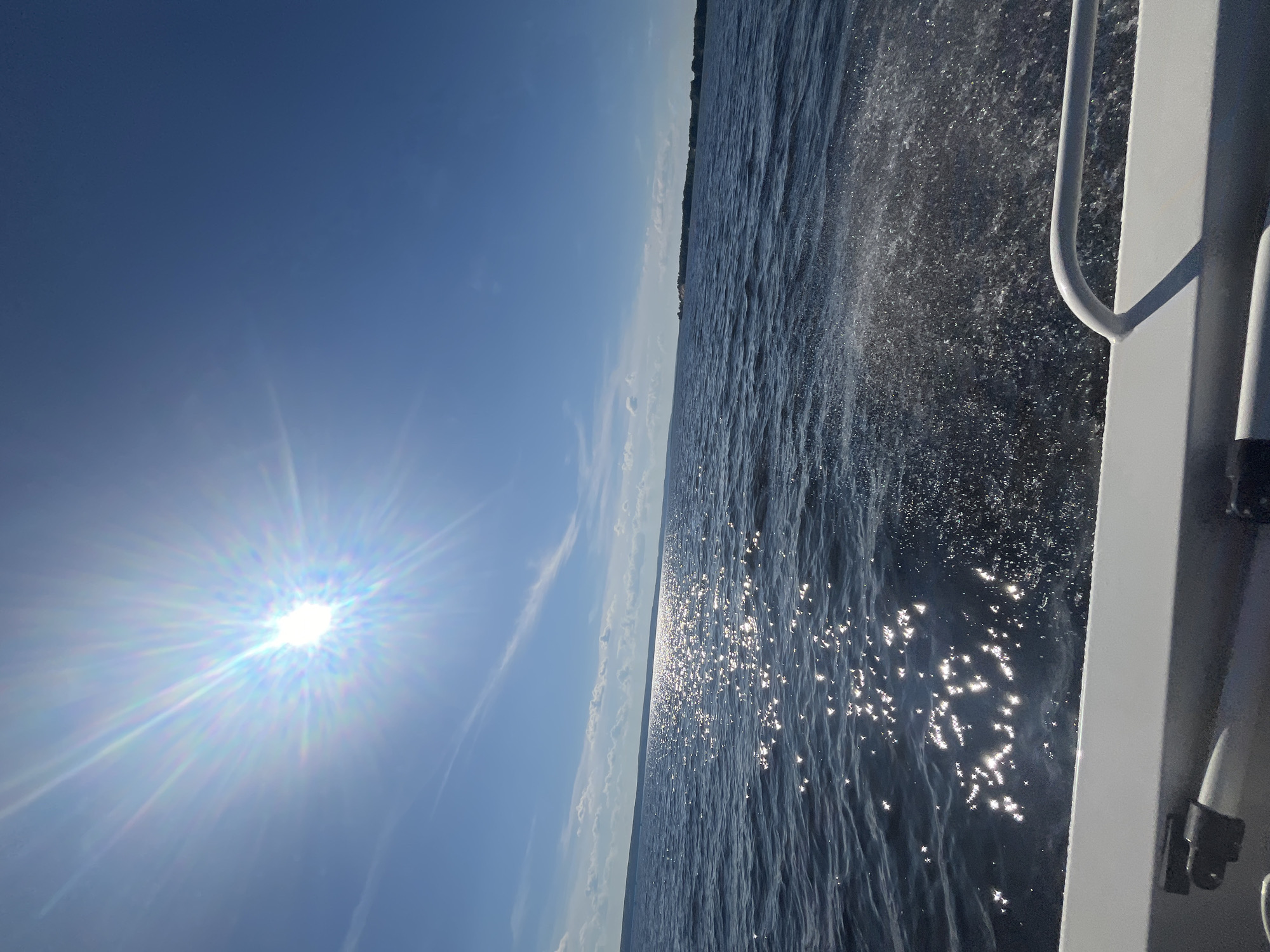 This media file captures a scenic view of a body of water, likely a lake or a wide river, under a bright, sunny sky. The sun, positioned in the upper left quadrant, is a brilliant starburst of light, casting a strong glare across the scene and creating shimmering reflections on the water's surface. wispy clouds streak across the otherwise clear blue sky.
The perspective suggests the image was taken from a moving vessel, possibly a boat. To the right of the frame, a portion of the boat's railing and superstructure is visible, rendered in a light color. The water is choppy, with spray being kicked up by the boat's movement, especially noticeable in the mid-right section of the image. The water itself is a deep, dark blue, contrasted by the bright, scattered highlights of sunlight.
In the distance, a faint, dark line of land is visible on the horizon, suggesting a shoreline or a distant landmass. The overall impression is one of a bright, clear day at sea or on a large lake, with a sense of movement and the enjoyment of being outdoors on the water. The time of day is clearly midday due to the intensity of the sun and the presence of strong shadows. There is no visible text in the media file.