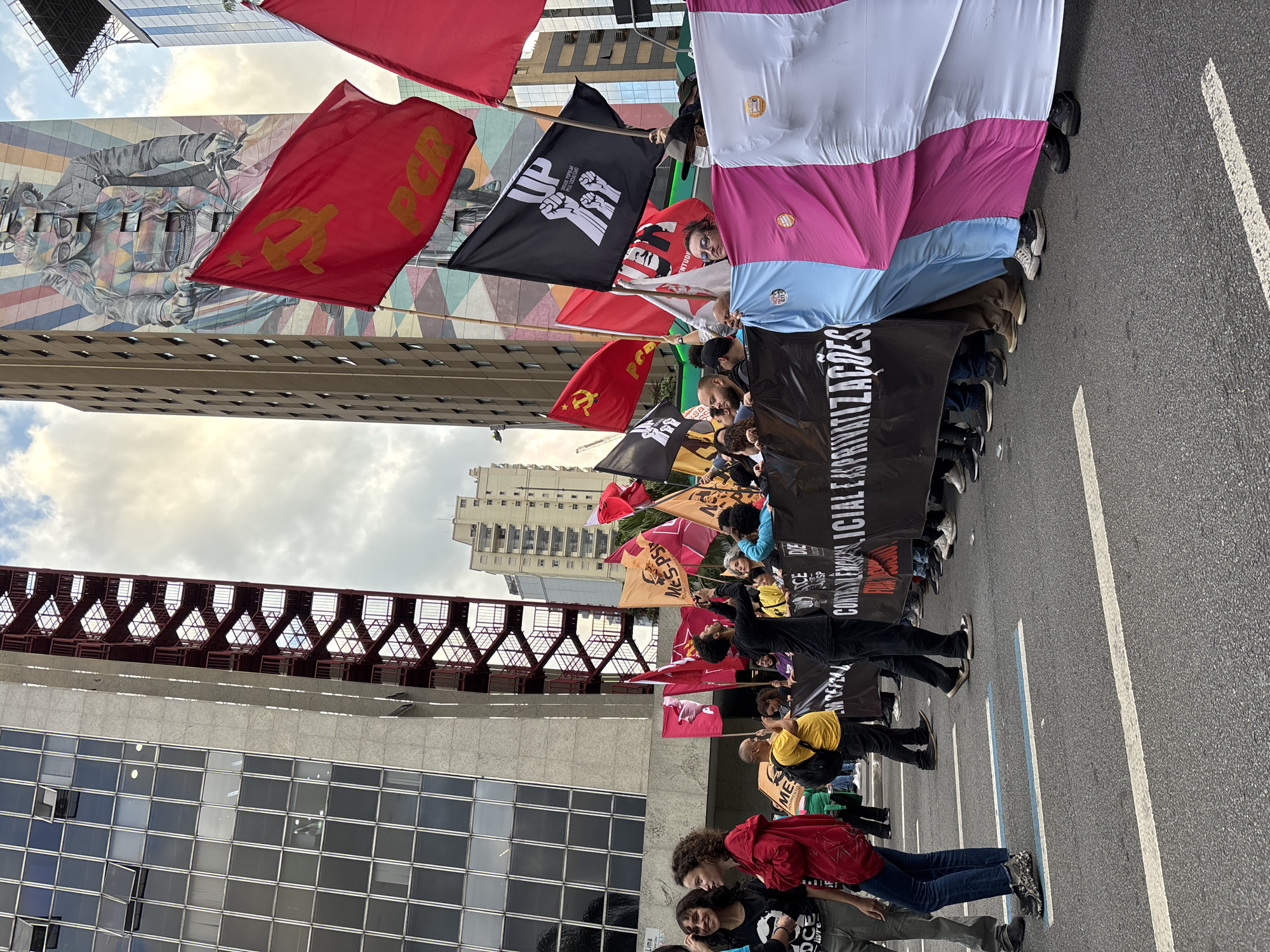 A crowd of people are demonstrating in the city of São Paulo, Brazil. They are carrying flags, including a red flag with a yellow hammer and sickle, a black flag with the words "UP" and "UNIDADE POPULAR" with three clenched fists, and a banner with Portuguese text "CONTRA A EXPLORAÇÃO SOCIAL E AS..." A mural is visible on a building in the background. The demonstration is taking place on a city street with buildings on both sides. A red, metal structure is also visible on the left side. The people are holding banners.
